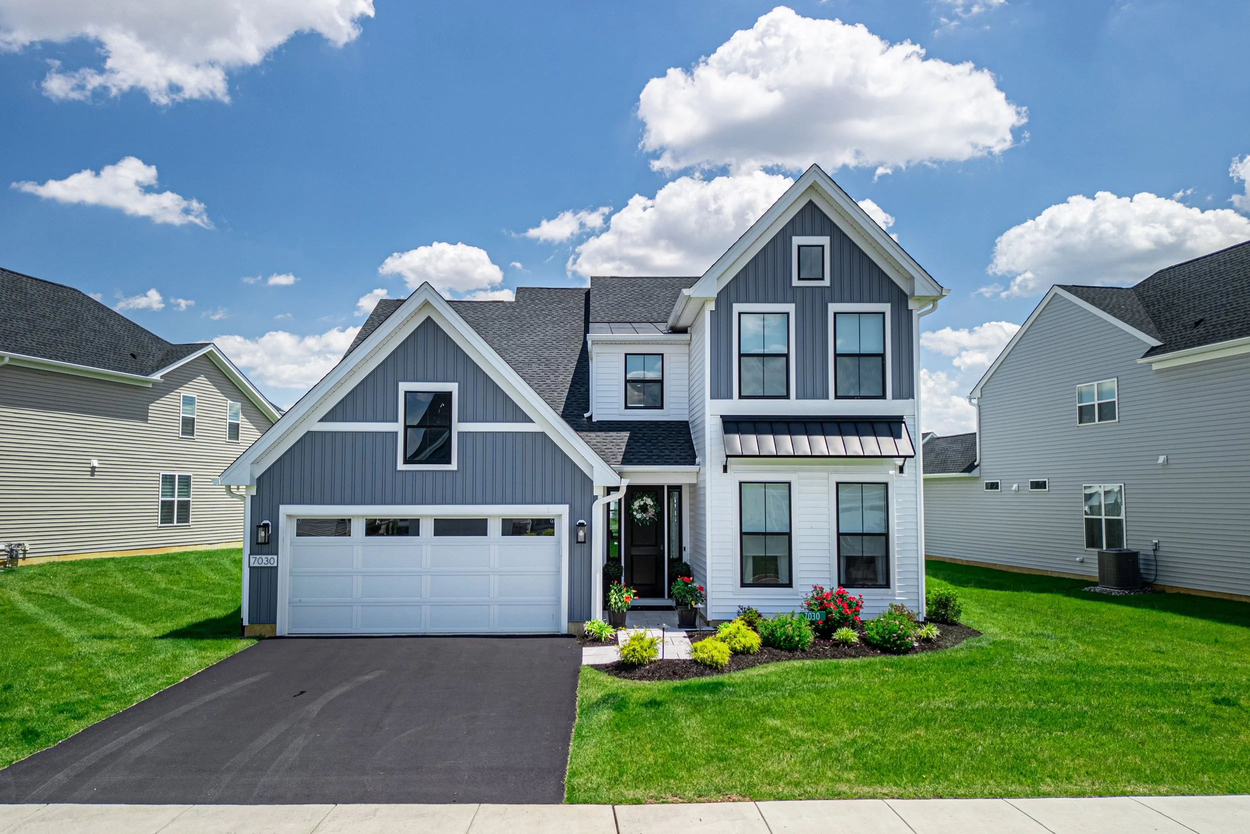 Modern two-story house with blue and white exterior, front yard with green grass and flower garden, driveway leading to a two-car garage, under a blue sky with scattered clouds.