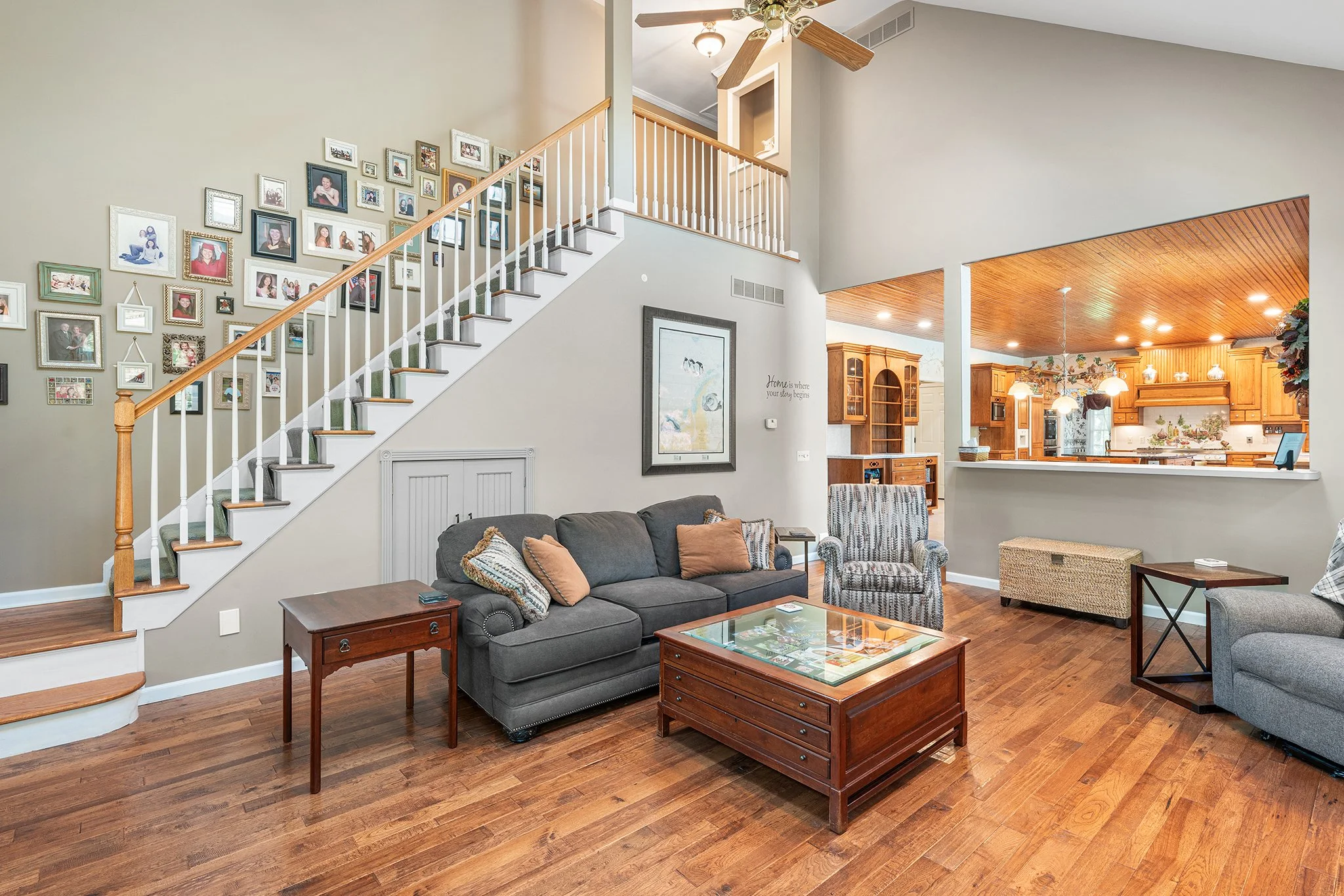 Living room with gray and patterned armchairs, wooden coffee table, dark gray sofa with pillows, wood floor, staircase with photo gallery, open kitchen with wooden cabinets, hanging lights, and ceiling fan.