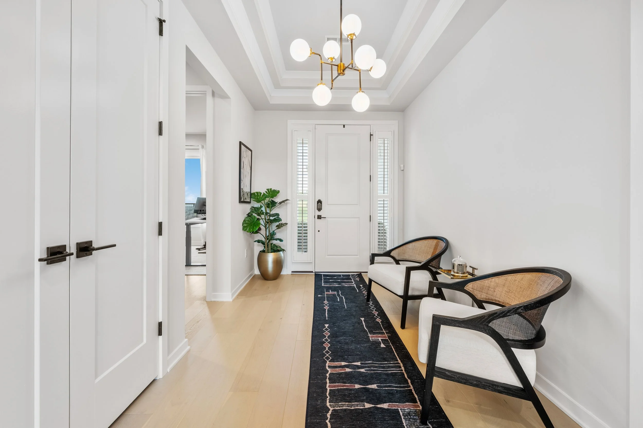 Modern entryway with white walls, a black and white rug, two white chairs with black frames, a tall green plant in a gold pot, and a gold chandelier with white globes.