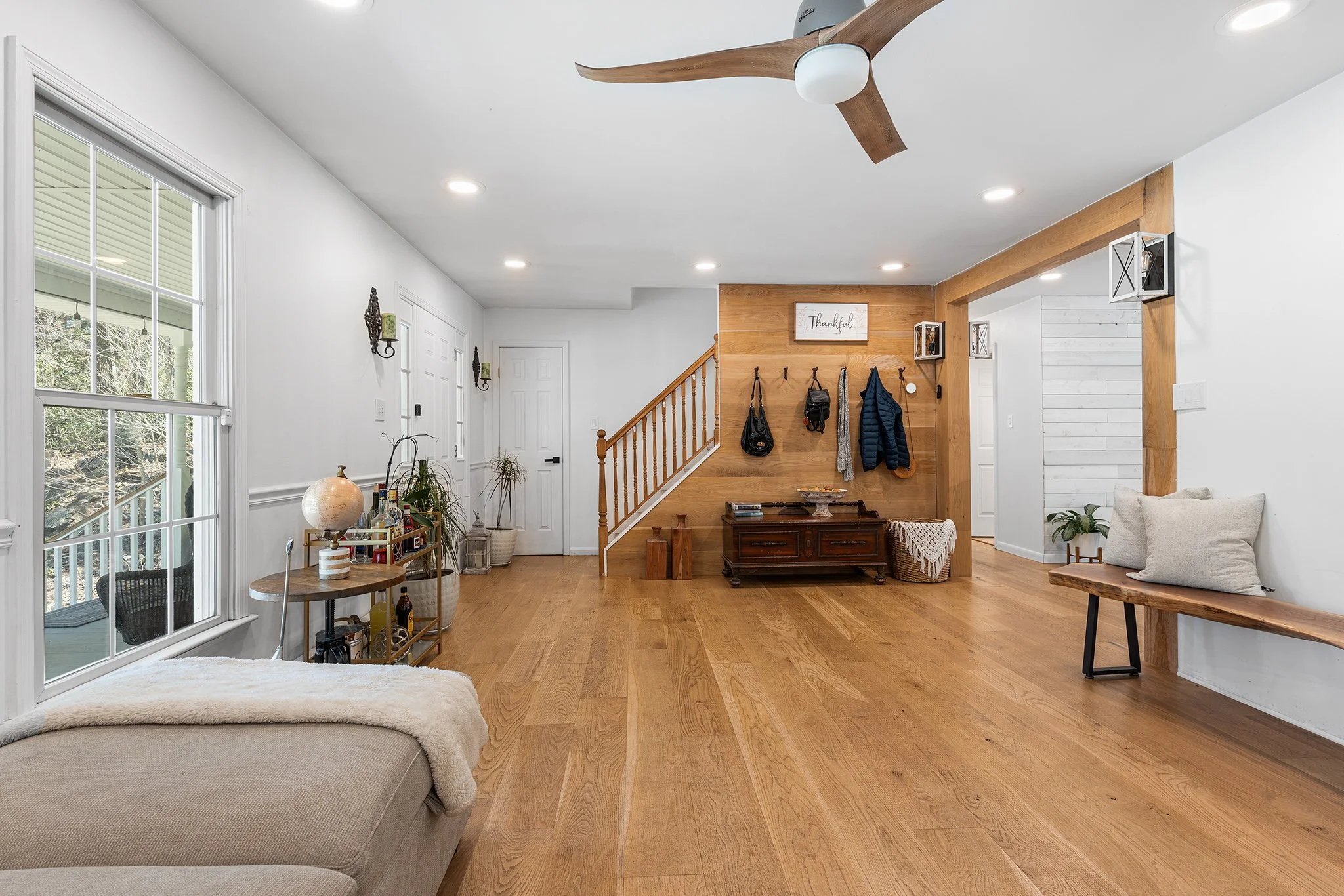 Living room with large window, wooden floors, wooden staircase, wall hooks with jackets, and minimal decor.