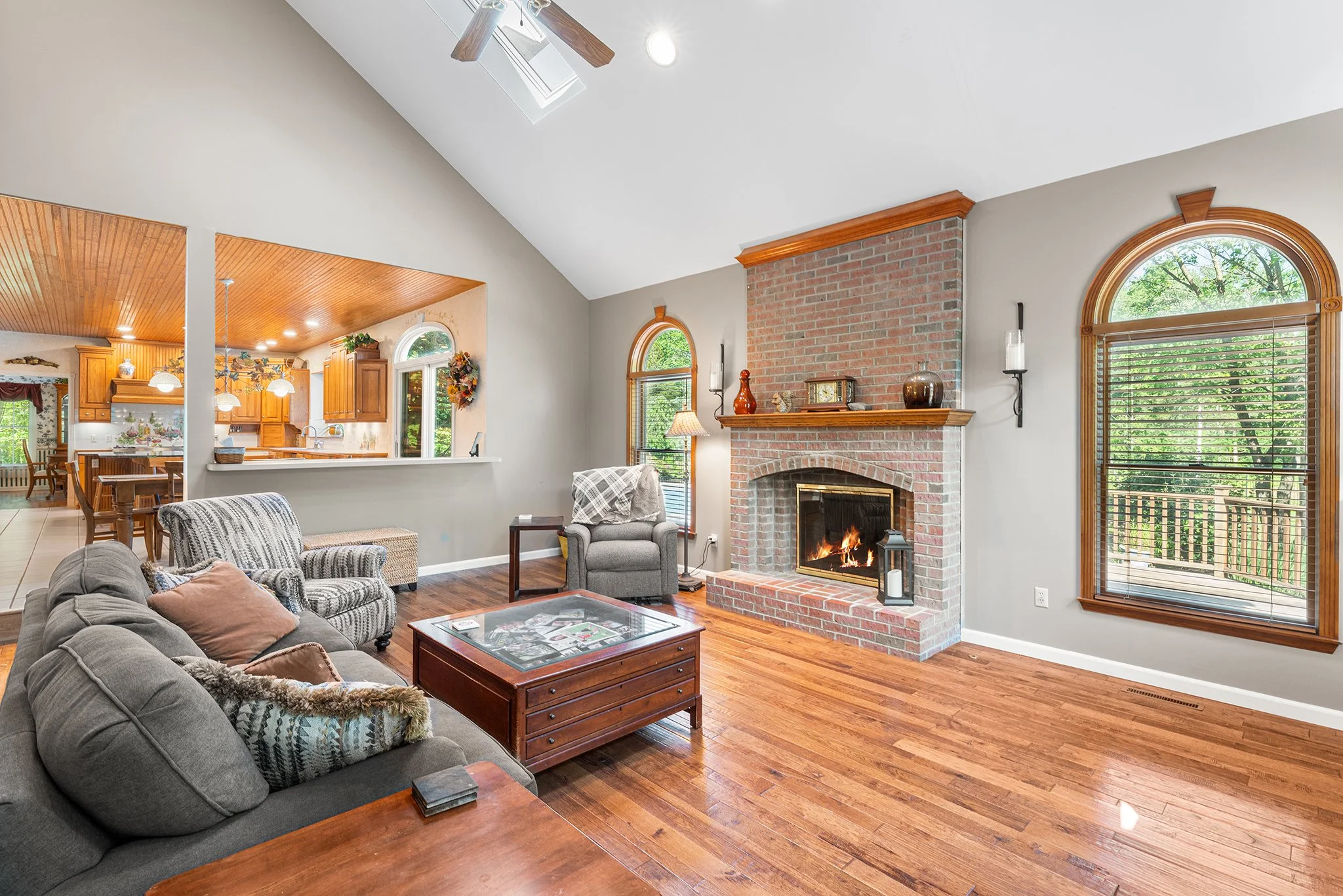 Living room with brick fireplace, arched windows, wooden floors, and a view into the kitchen.