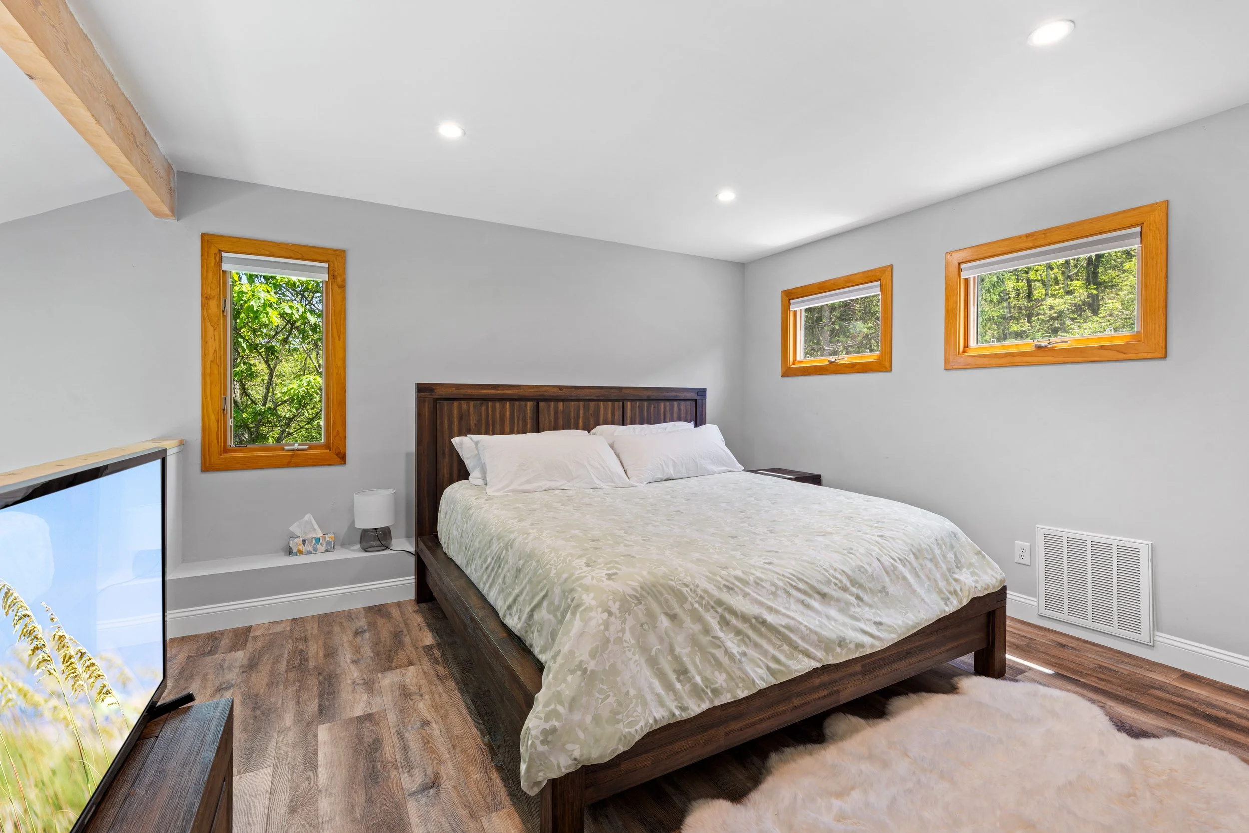 A bedroom with white walls, three windows with wooden frames, a wooden bed frame with a light-colored blanket, a small bedside table, a white lamp, and a flat-screen TV on a wooden stand, with a fluffy rug on the floor.