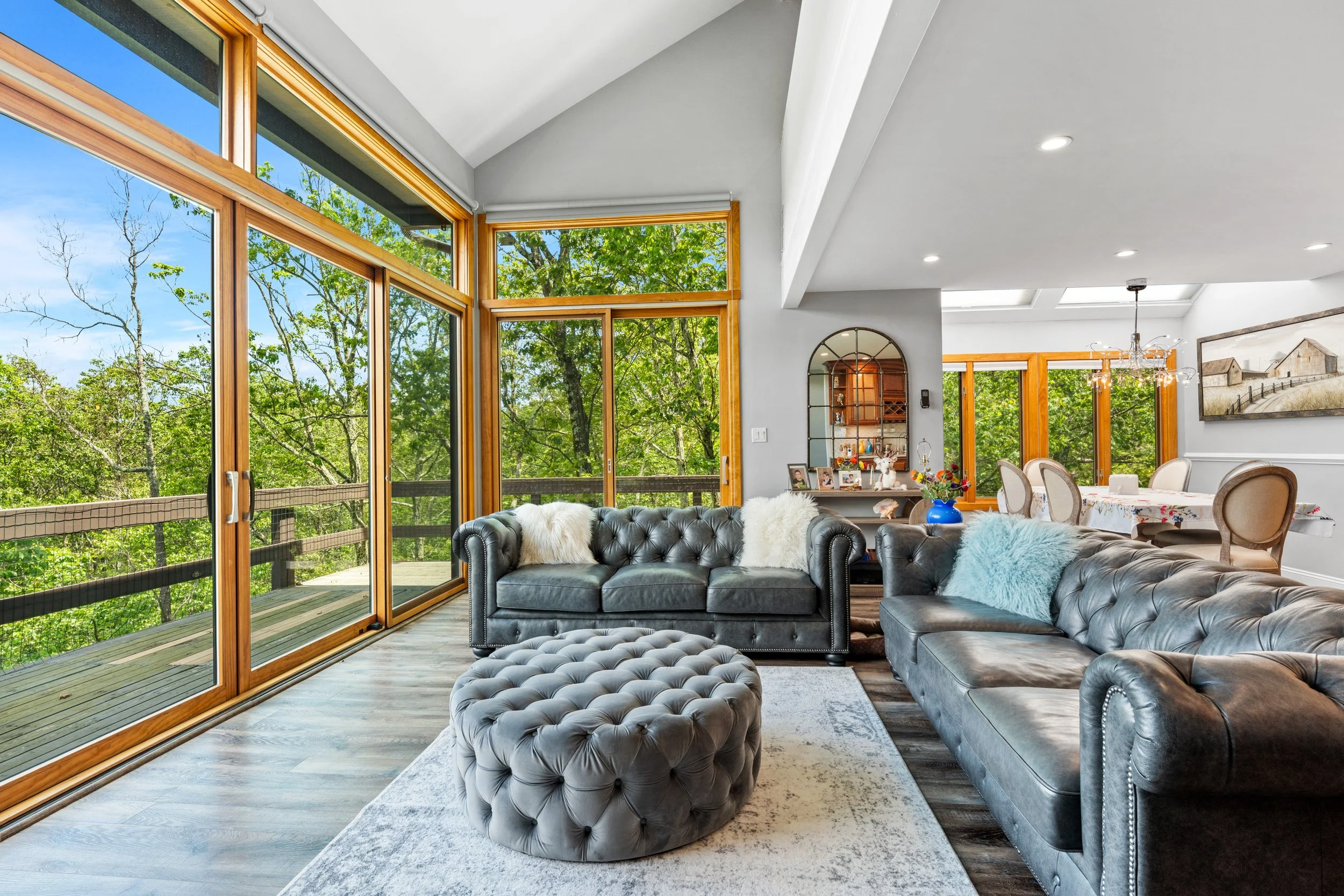 Living room with large windows and a view of green trees, featuring black leather sofas, a tufted ottoman, and a dining area in the background.
