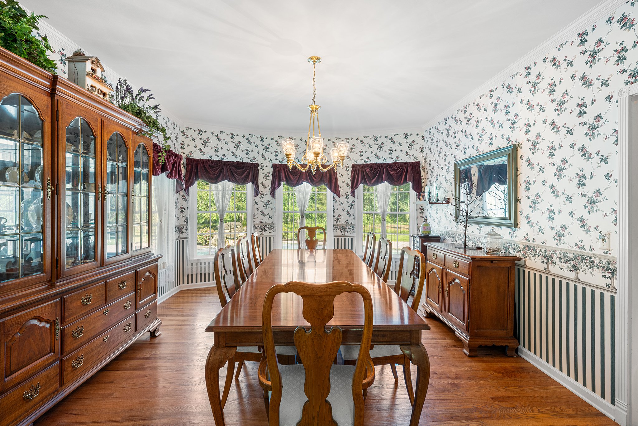 A traditional dining room with a long wooden table and eight matching chairs, floral patterned wallpaper, three windows with white curtains and burgundy valances, a chandelier hanging from the ceiling, a wooden china cabinet on the left, and a sidebo