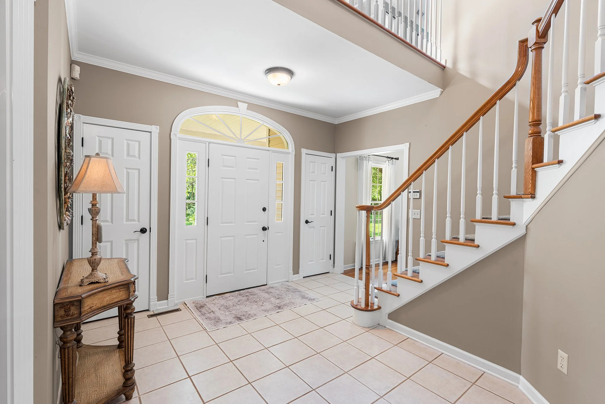 Entryway with tiled floor, white front door with sidelights, staircase with wooden handrail, beige walls, window with curtains, and a small wooden table with a lamp.