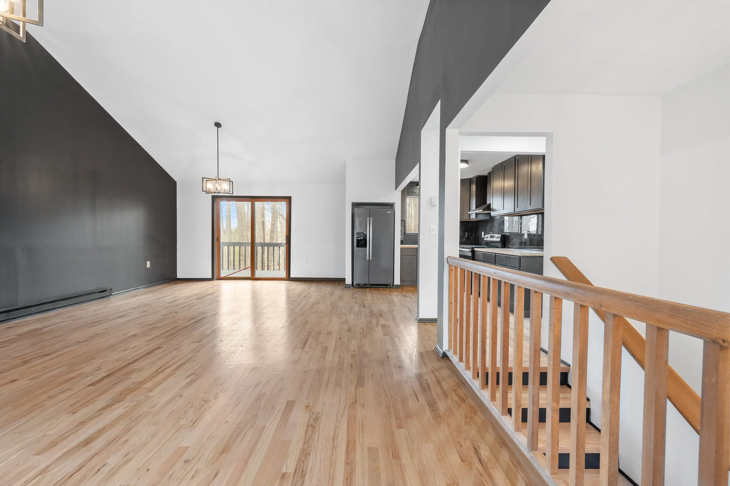 Empty living room with hardwood floors, black accent wall, and staircase with wooden railing leading downstairs. Kitchen with dark cabinets and stainless steel refrigerator visible through an open wall. Sliding glass door opens to a balcony or porch.