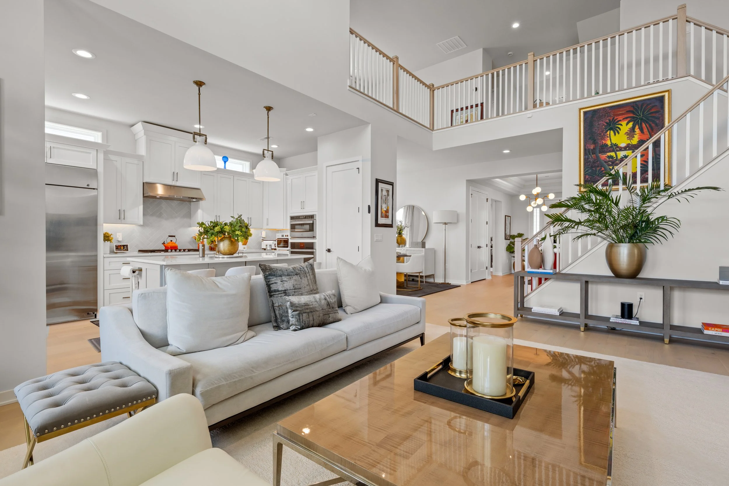 Open-concept living room with a white sofa, a wooden coffee table with candles, and a staircase with a large palm plant and colorful sunset painting. The background shows a modern kitchen with white cabinets, pendant lights, and a small dining area.