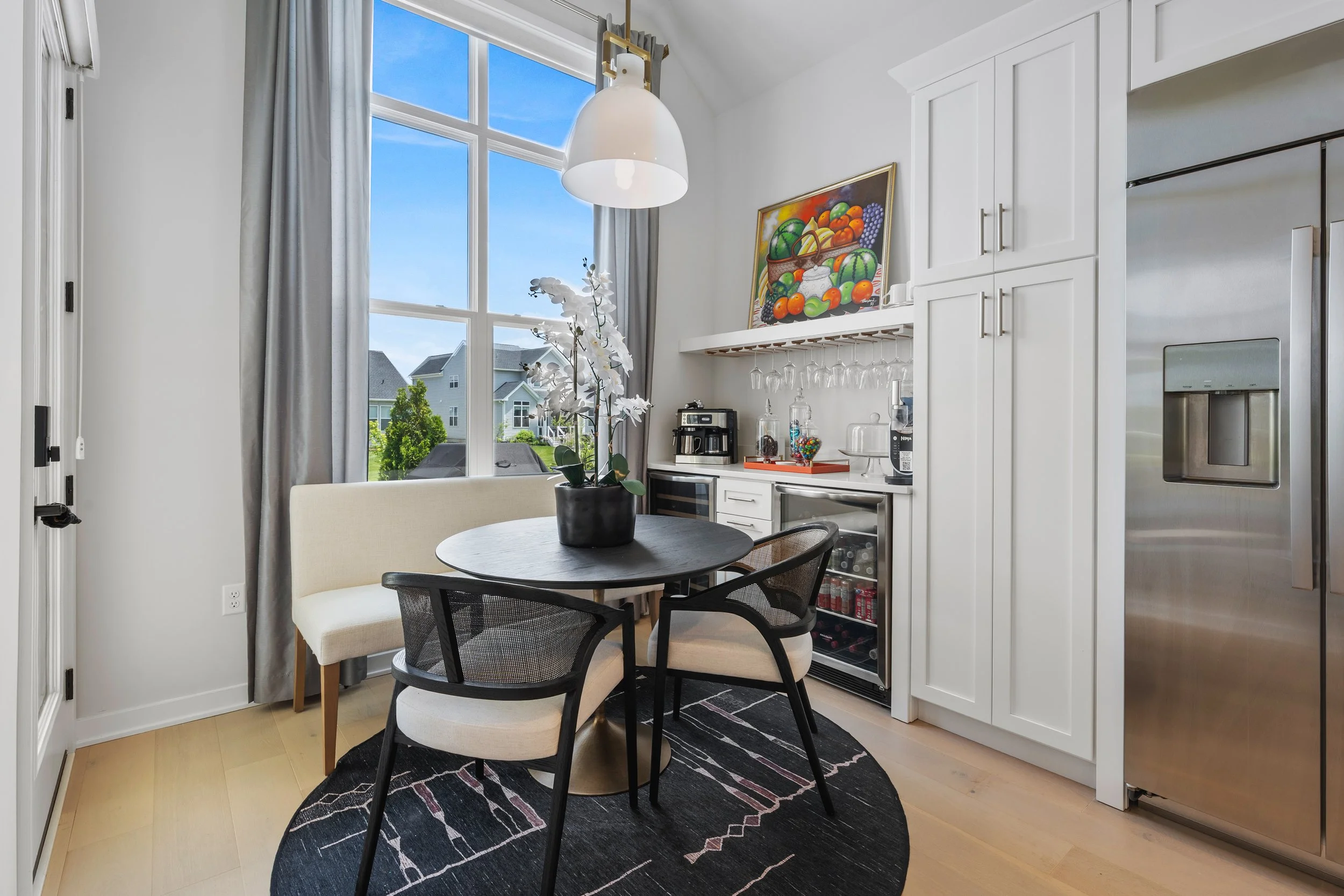 Kitchen corner with round black table, four chairs, a white cushioned bench, potted orchid, large window with gray curtains, white cabinetry, and a stainless steel refrigerator.