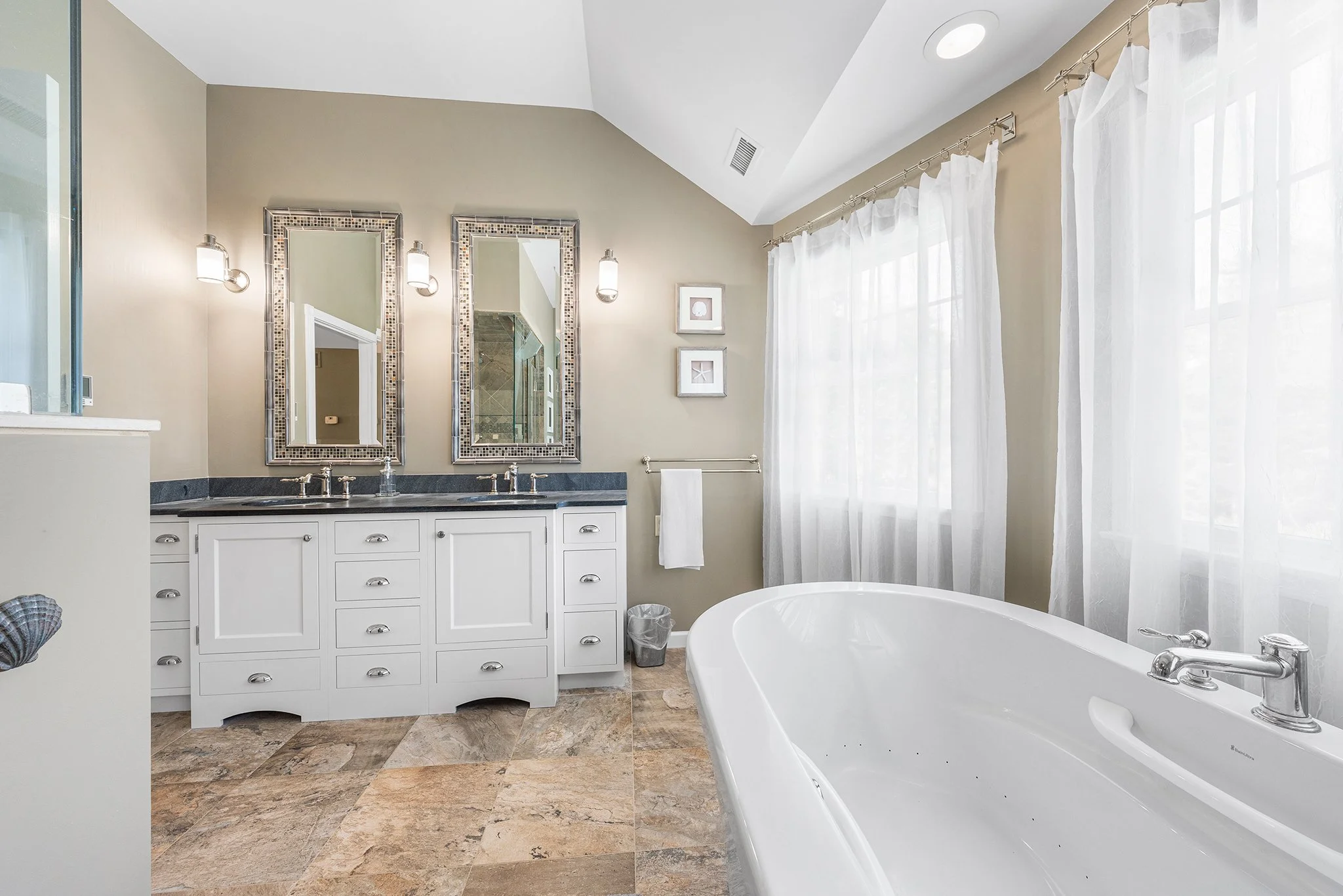 Bright bathroom with double vanity, two mirrors, and a large bathtub under windows with sheer white curtains.