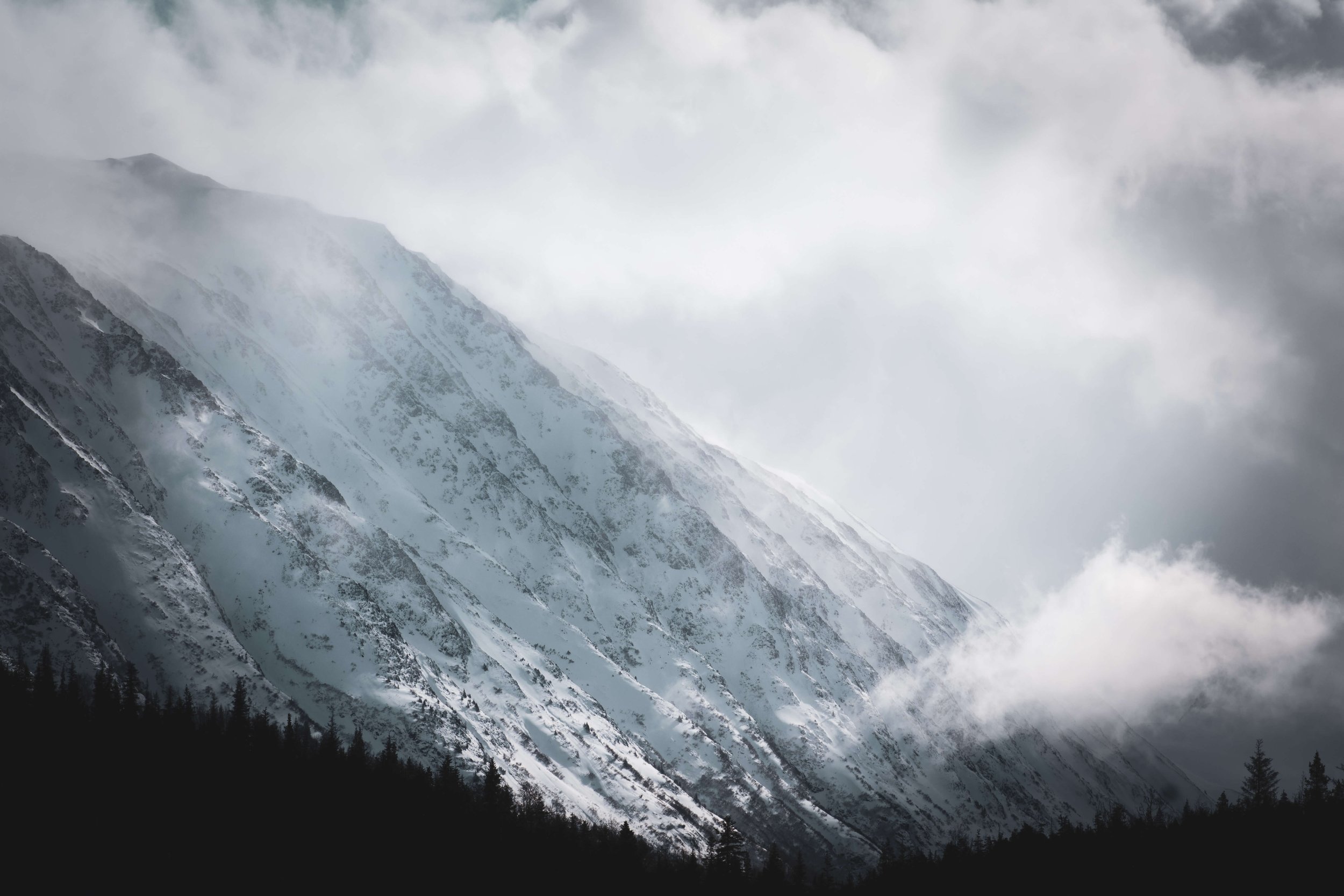 photograph of snowcapped mountain peaks face, intimidating mountain range, sun shining above, soft lighting across the mountains, clouds, silhouette of a forest, taken in the Alaskan wilderness, Alaska, USA.