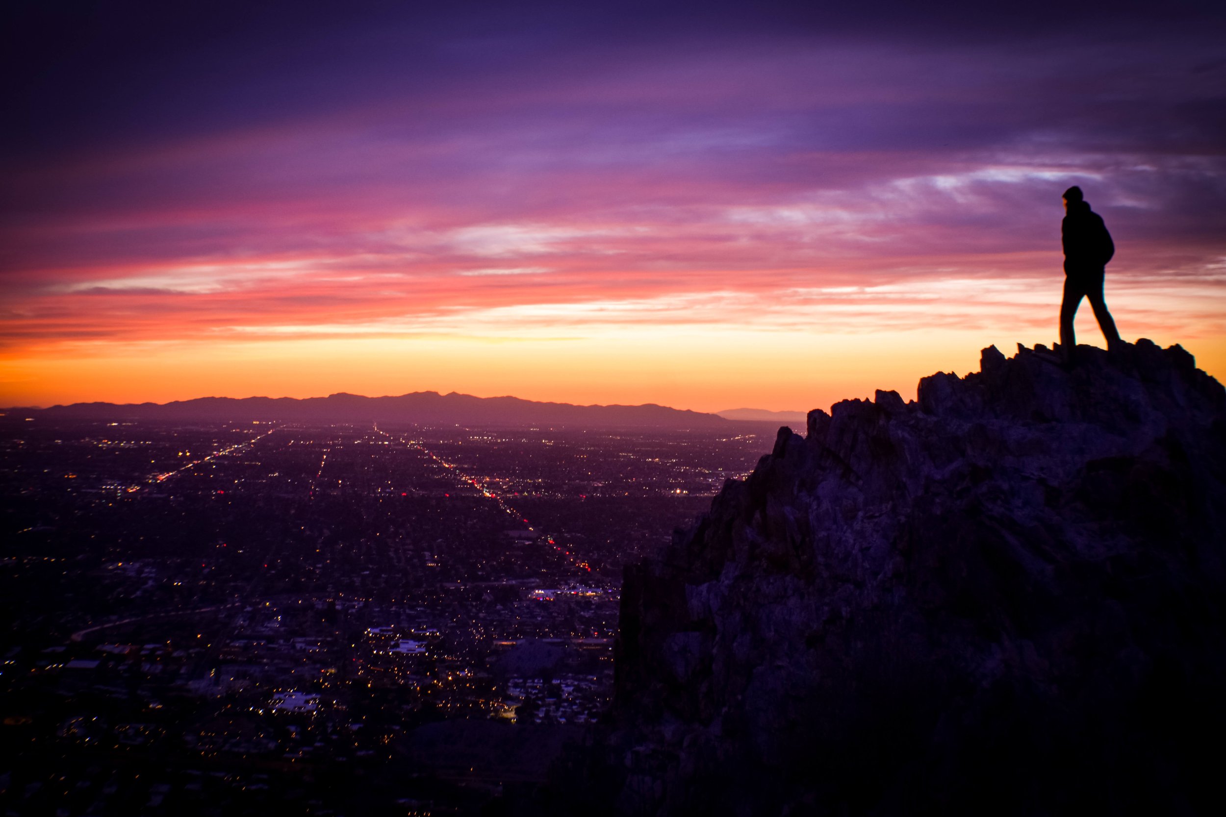 The silhouette of a lone man stands on the top of Piestewa Mountain Peak at sunset, the city of Phoenix, Arizona, USA is illuminated in evening city lights below, hiking, hiking trail, Maricopa, sunset desert sky, Phoenix mountains, Squaw Peak, Camel