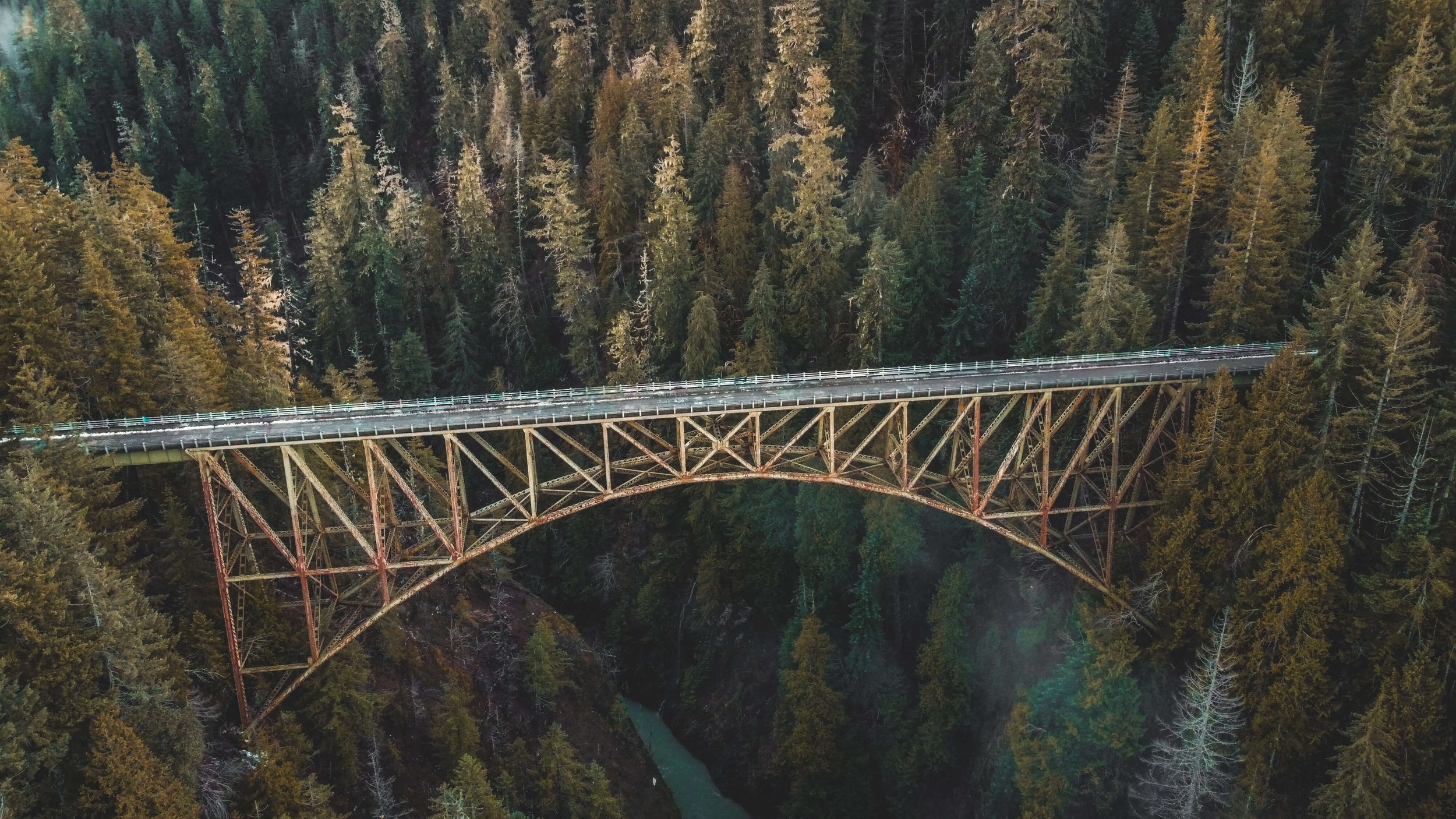 a drone photograph taken from the air of a bridge over a glacial blue winding river, an green evergreen forest in Washington state, Pacific Northwest, USA, high steel bridge, Skokomish River.