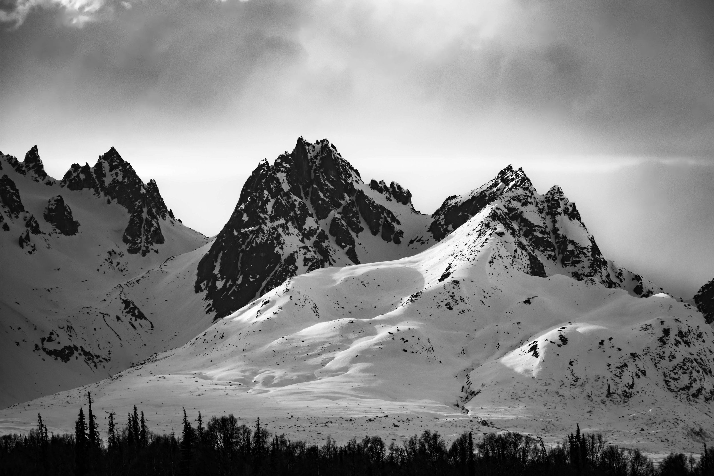 black and white photograph of snowcapped mountain peak face, intimidating mountain range, sun shining above, soft lighting across the mountains, taken in the Alaskan wilderness, Alaska, USA.