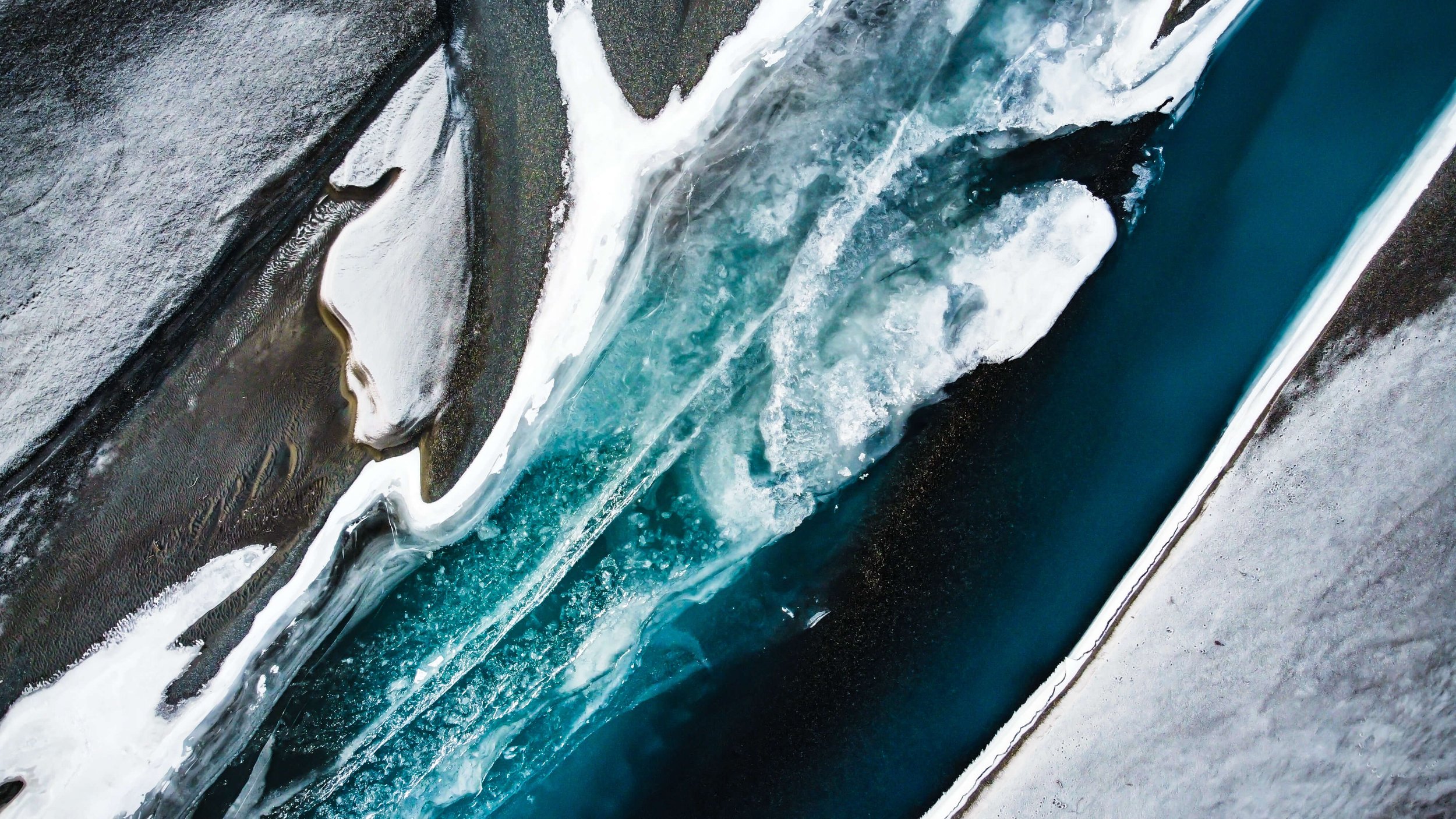 a drone photograph taken from the air of a frozen winding river in Alaska wilderness, abstract nature photography, glacial blue, white ice, black sand and sediments, Alaska, USA.