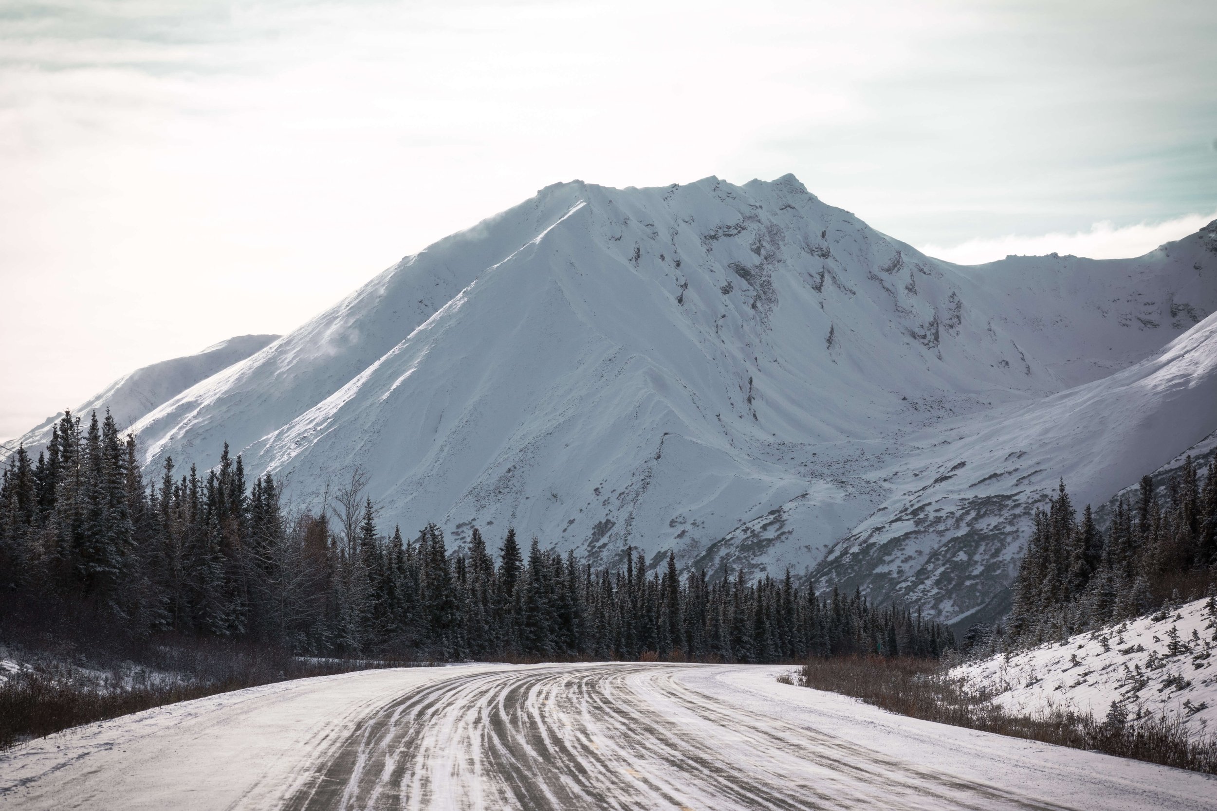 black and white photograph of a remote snow ice covered highways, snowcapped mountain peak face, intimidating mountain range, sun shining above, taken in the Alaskan wilderness, Alaska, USA, road shot, winter highway.