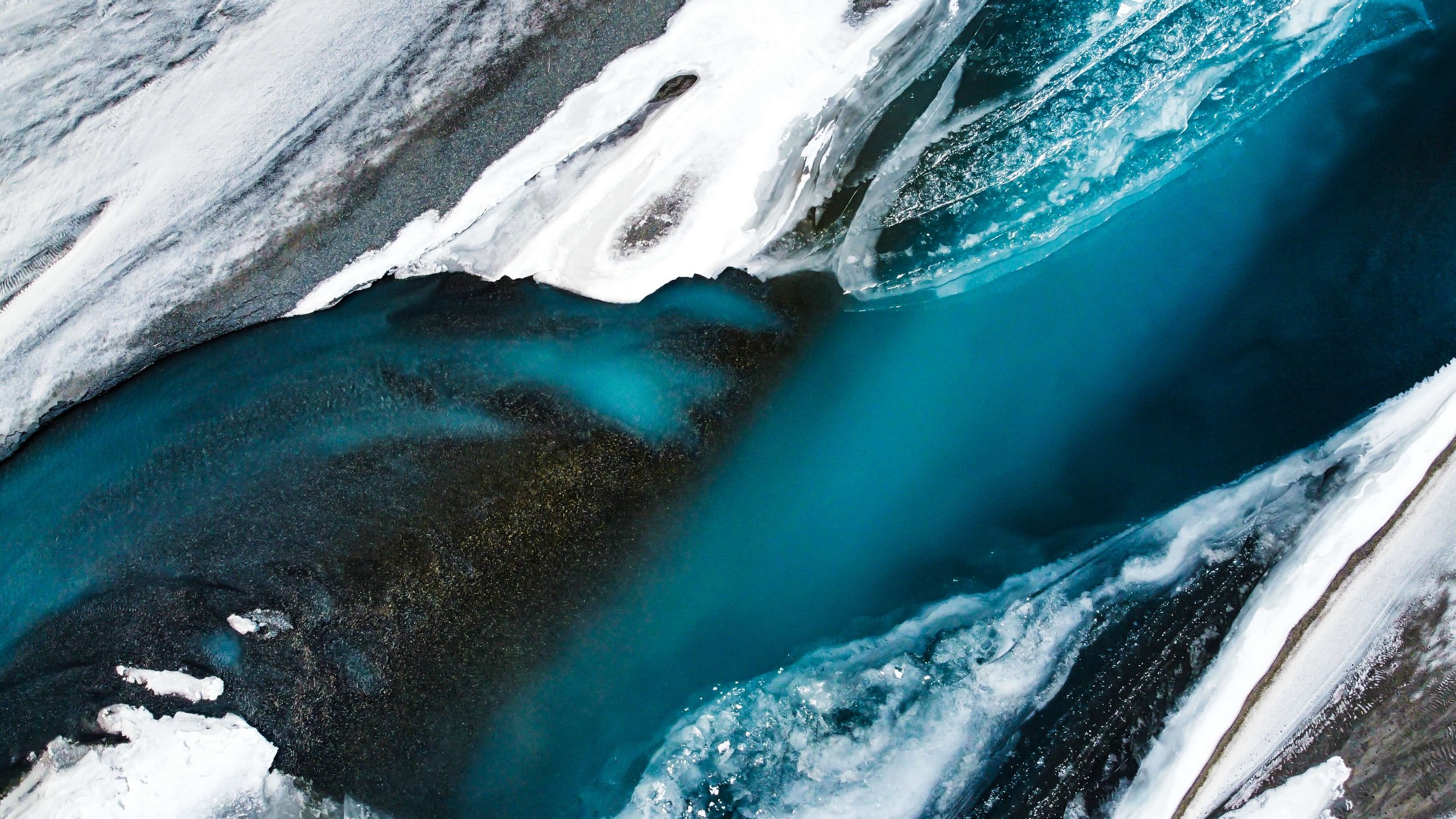 a drone photograph taken from the air of a frozen winding river in Alaska wilderness, abstract nature photography, glacial blue, white ice, black sand and sediments, Alaska, USA.