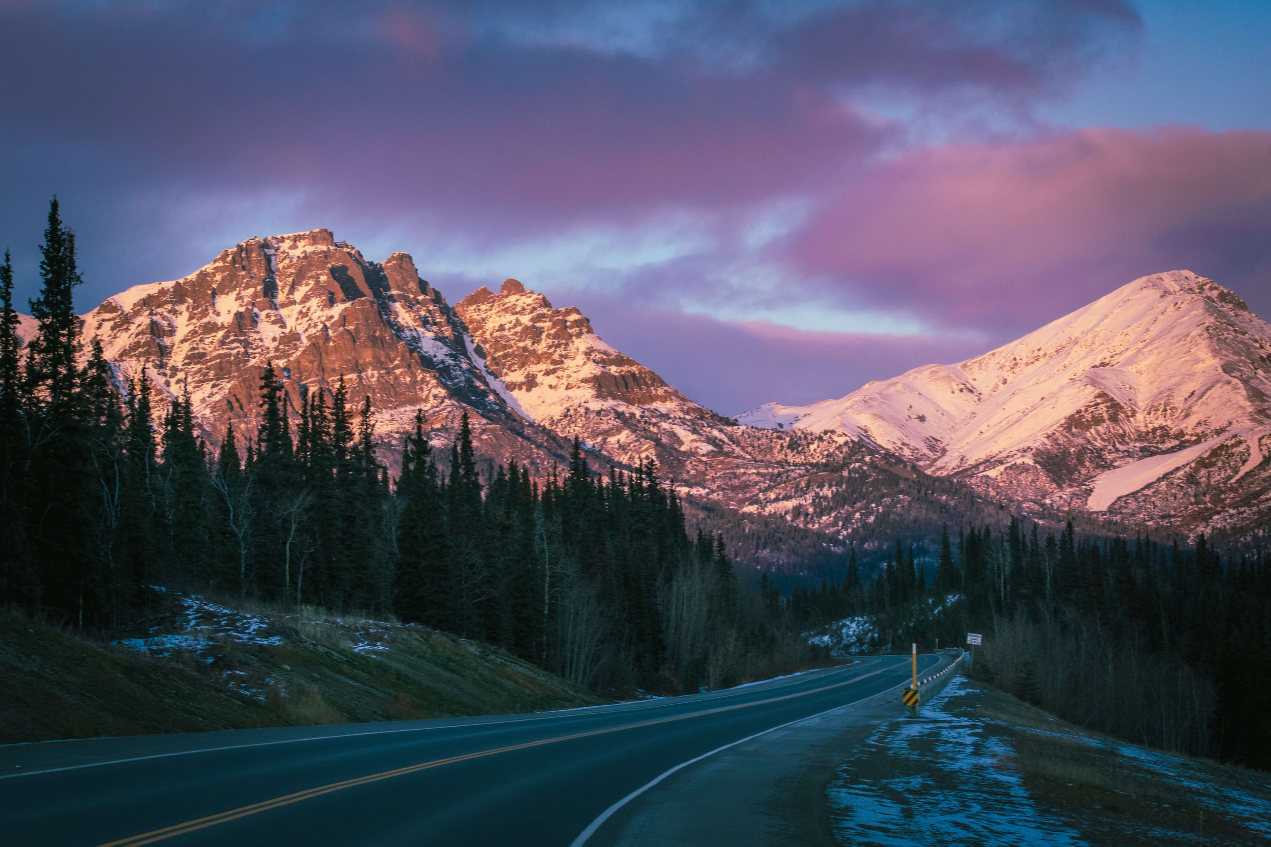 a remote lone highway in the Alaskan wilderness, Alaska, USA, snowcapped mountain peaks, intimidating mountain range, sunrise, sunset sun shining above, soft blue and pink lighting across the mountains, pink clouds, forest trees in the foreground, ro