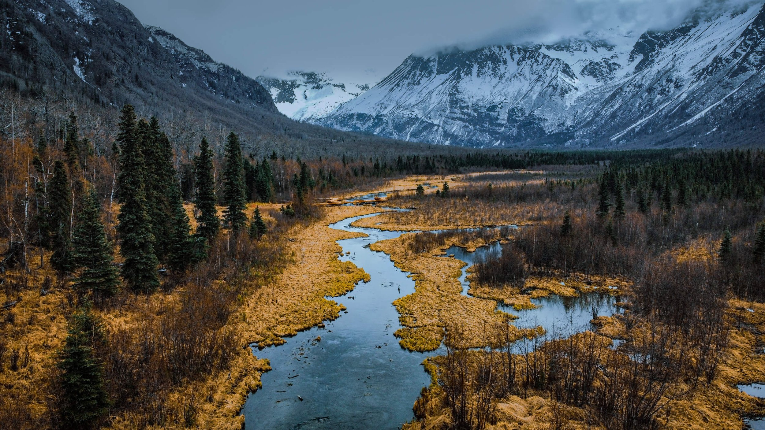a drone photograph taken from the air of a glacial blue winding river cutting through a frozen meadow in the remote Alaska wilderness, frozen pines, snowcapped mountain peaks, Alaksa, USA, Seaward highway, Anchorage.