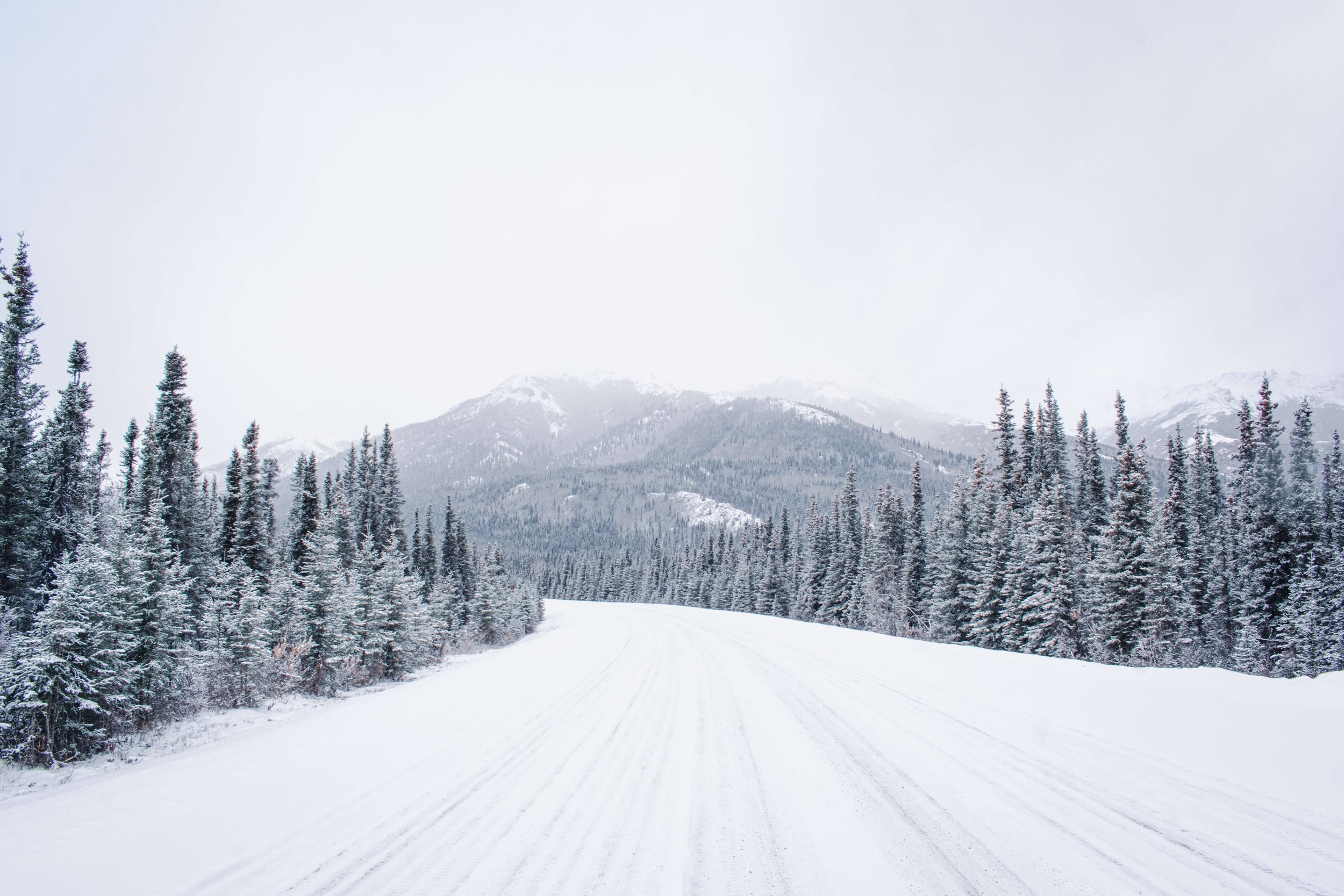 black and white photograph of a remote snow ice covered highway road, snowcapped mountain peak face, intimidating mountain range, taken in the Alaskan wilderness, Alaska, USA, Denali National Park, winter highway, road shot.