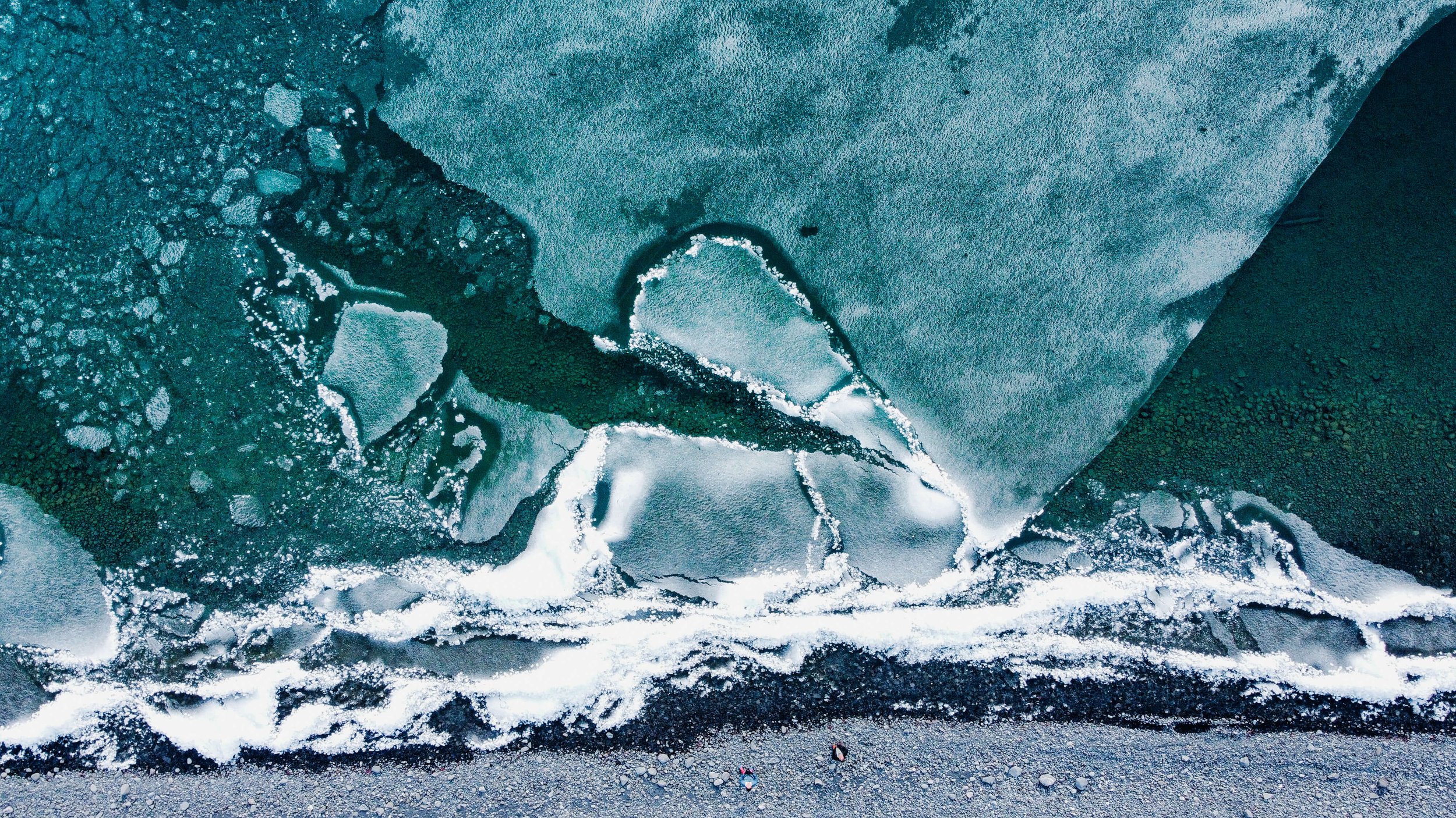 a drone photograph taken from the air of two male men standing at the edge of a frozen lake in remote Alaska wilderness, abstract nature photography, glacial blue, white ice, rocky shoreline and sediments, Alaska, USA.