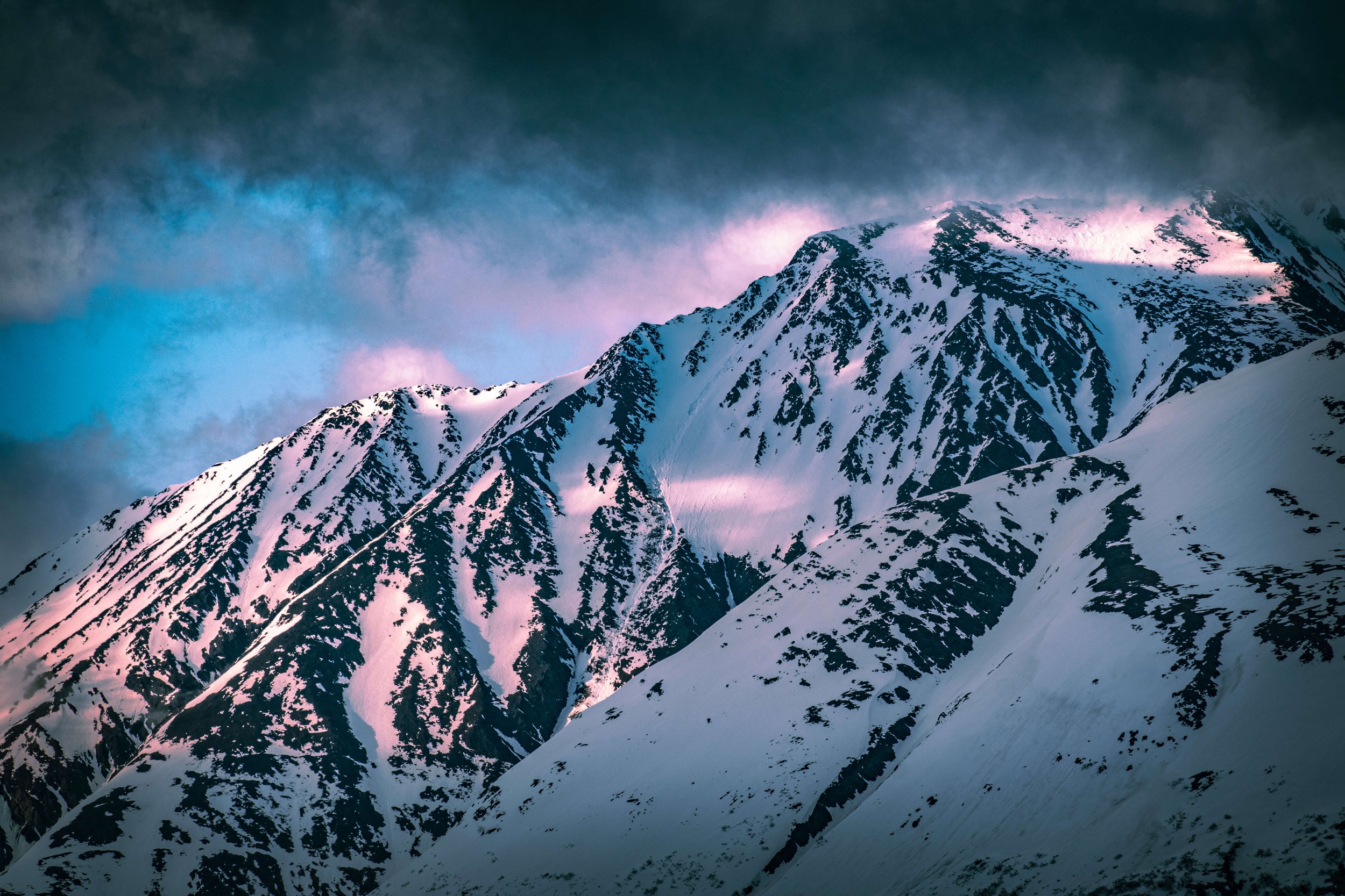 photograph of snowcapped mountain peaks face, intimidating mountain range, sunrise, sunset sun shining above, soft blue and pink lighting across the mountains, pink clouds, taken in the Alaskan wilderness, Alaska, USA.