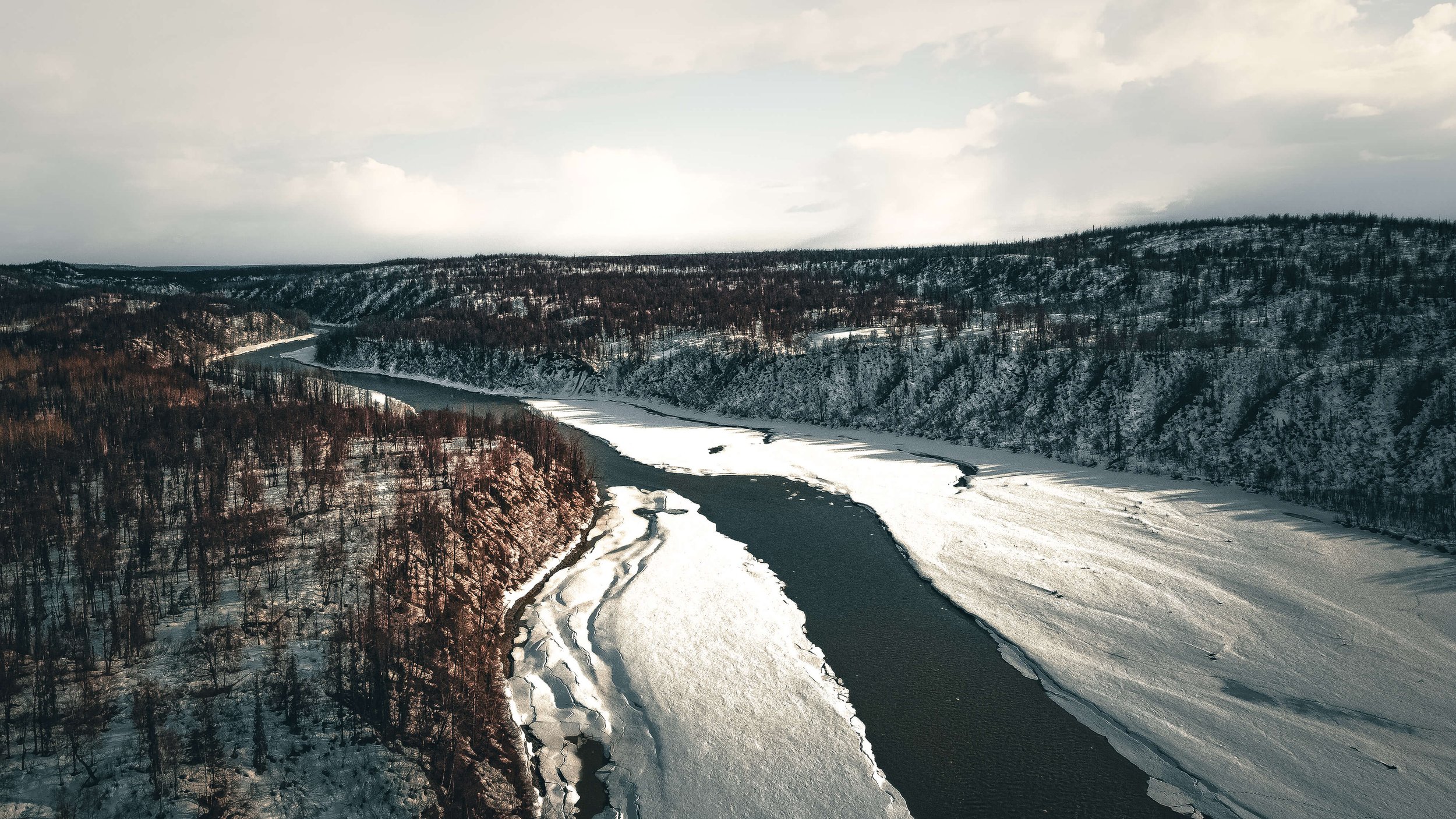 a drone photograph taken from the air of a frozen winding river in Alaska wilderness, soft lighting, frozen pine forest, Alaska, USA.