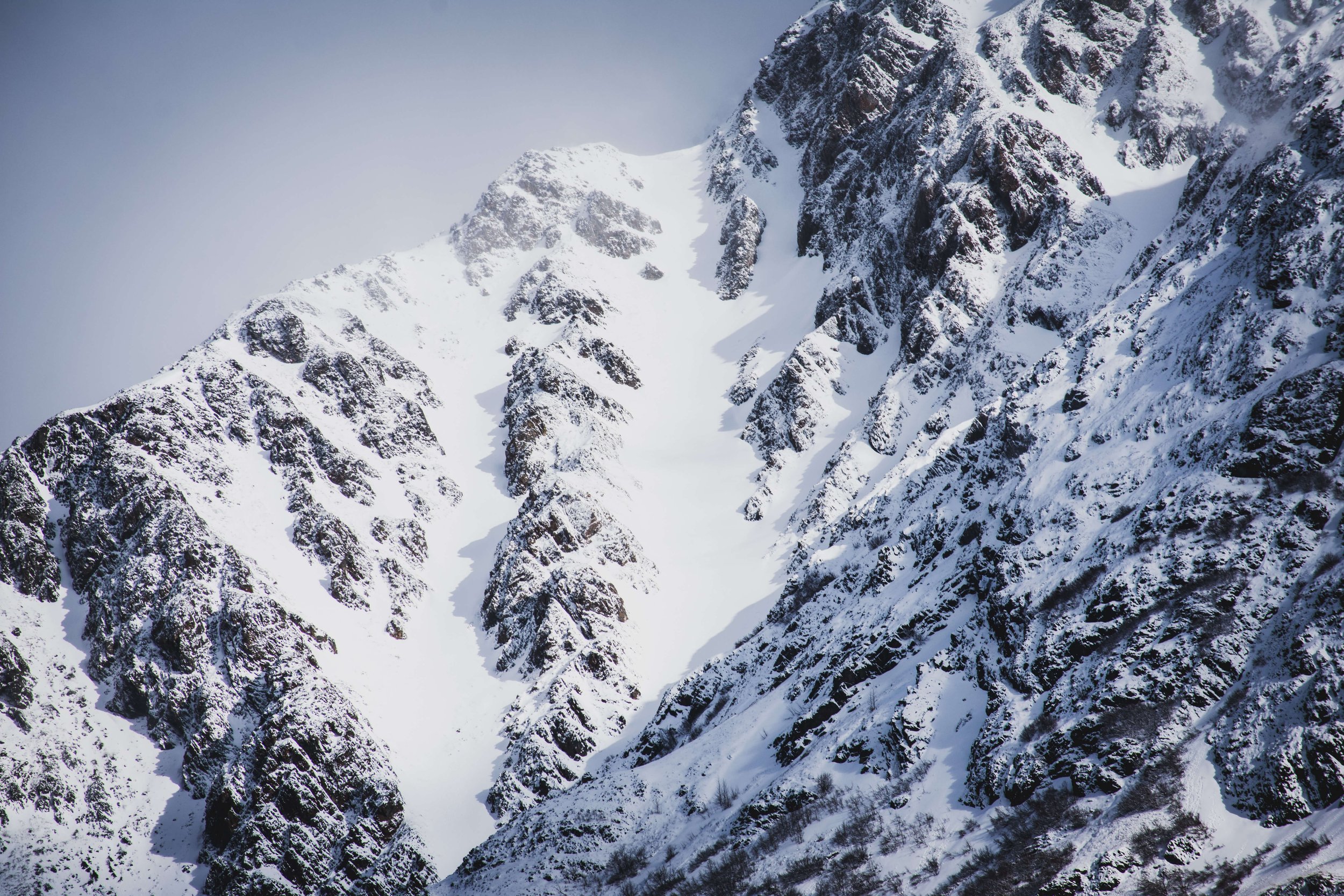 black and white photograph of snowcapped mountain peak face, intimidating mountain range, sun shining above, soft lighting across the mountains, taken in the Alaskan wilderness, Alaska, USA.