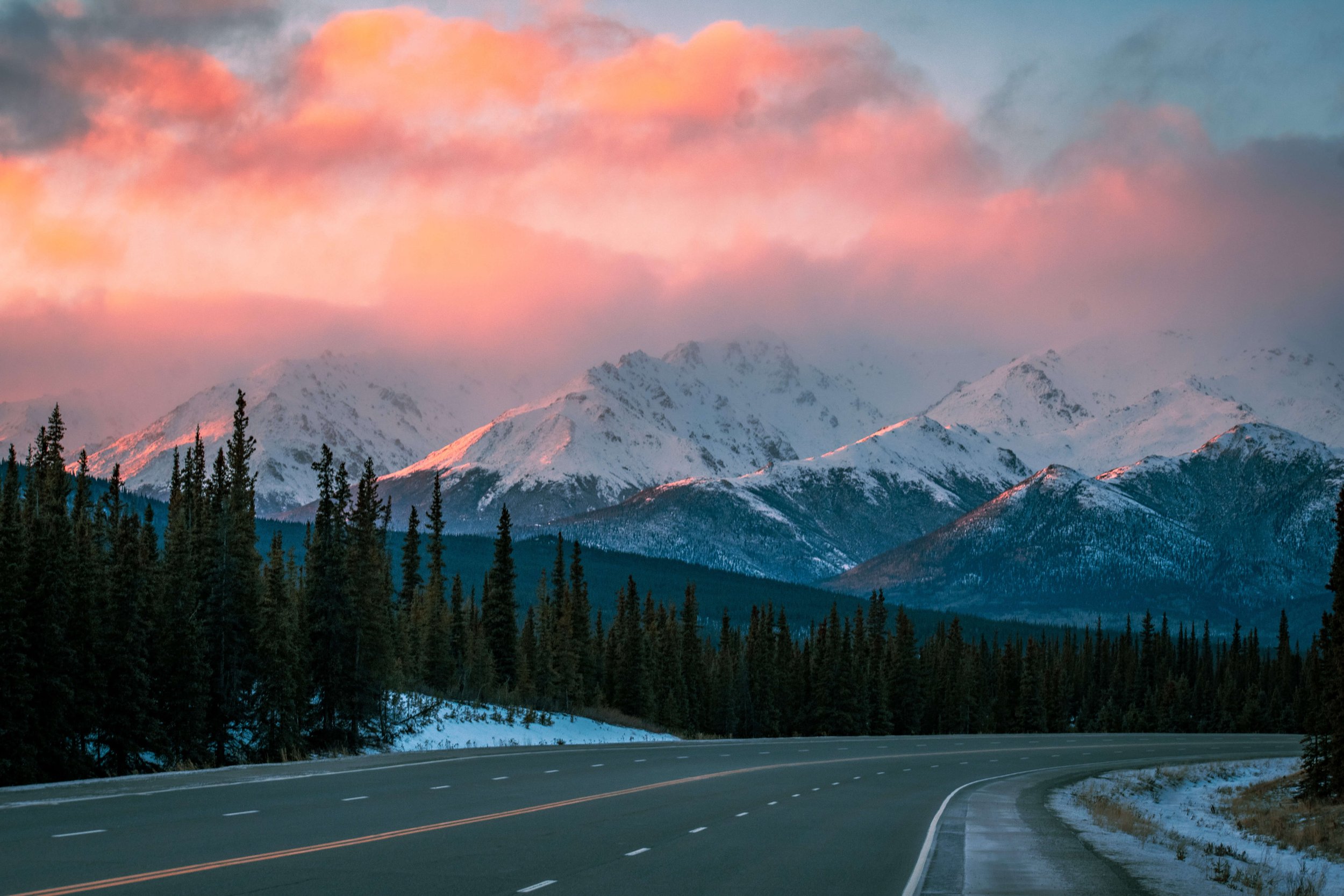 a remote lone highway in the Alaskan wilderness, Alaska, USA, snowcapped mountain peaks, intimidating mountain range, sunrise, sunset sun shining above, soft blue and pink lighting across the mountains, pink clouds, forest trees in the foreground, ro