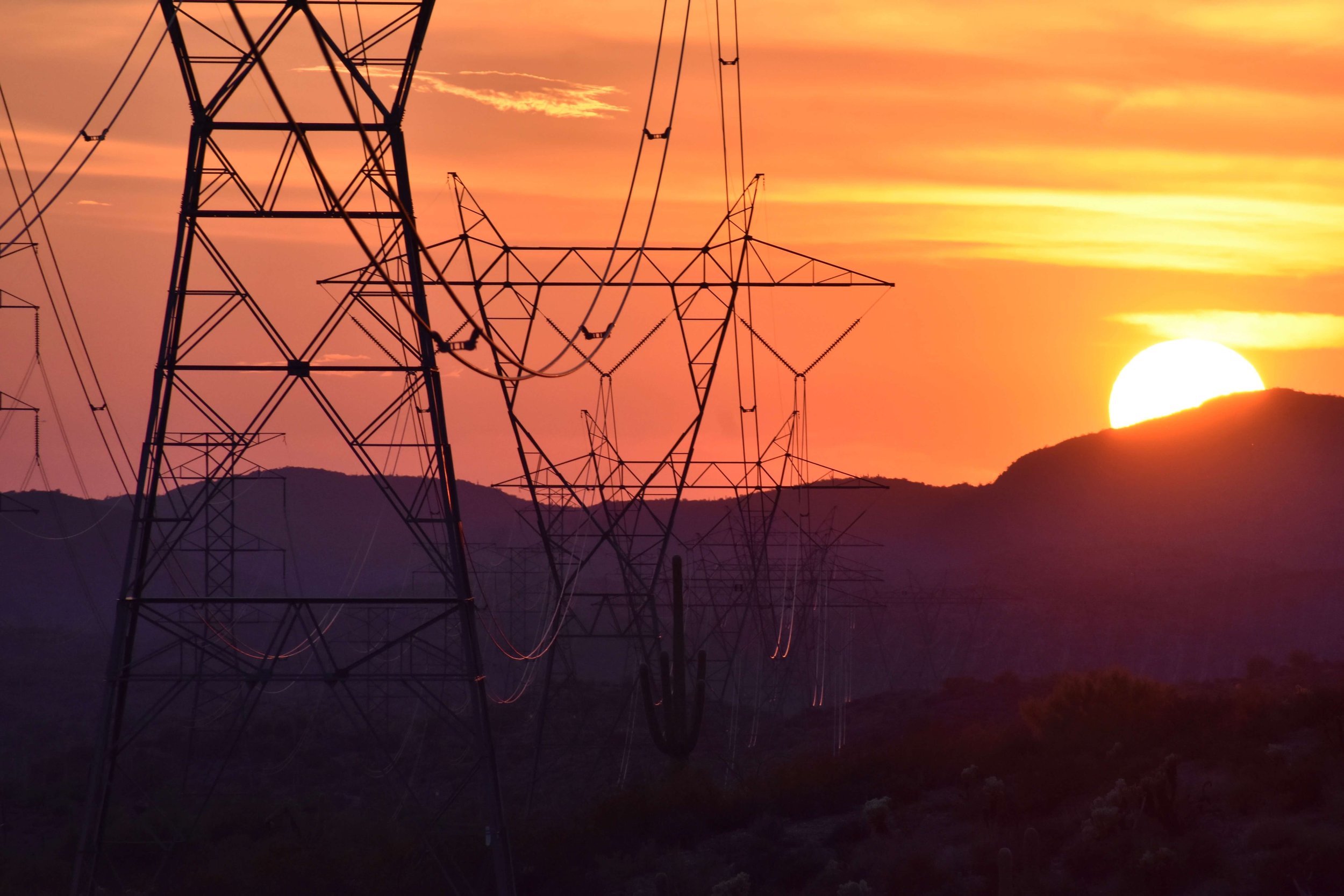 An image of the sun setting behind a desert mountain, creating a sky colored in fiery red, crimson, maroon, and orange, silhouettes of power lines extending into the distance, sunset desertscape, Arizona, USA, saguaro cactus, golden hour, American So