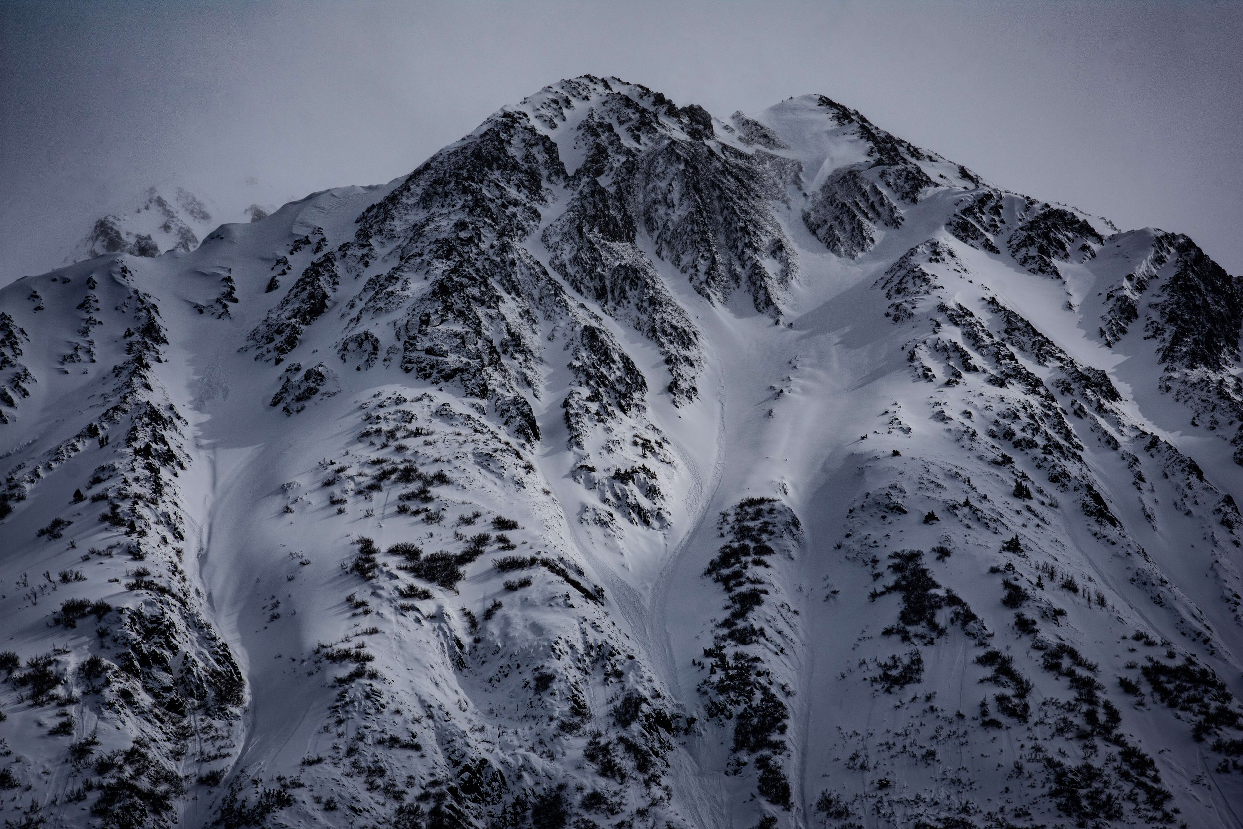 black and white photograph of snowcapped mountain peak face, intimidating mountain range, sun shining above, soft lighting across the mountains, taken in the Alaskan wilderness, Alaska, USA.