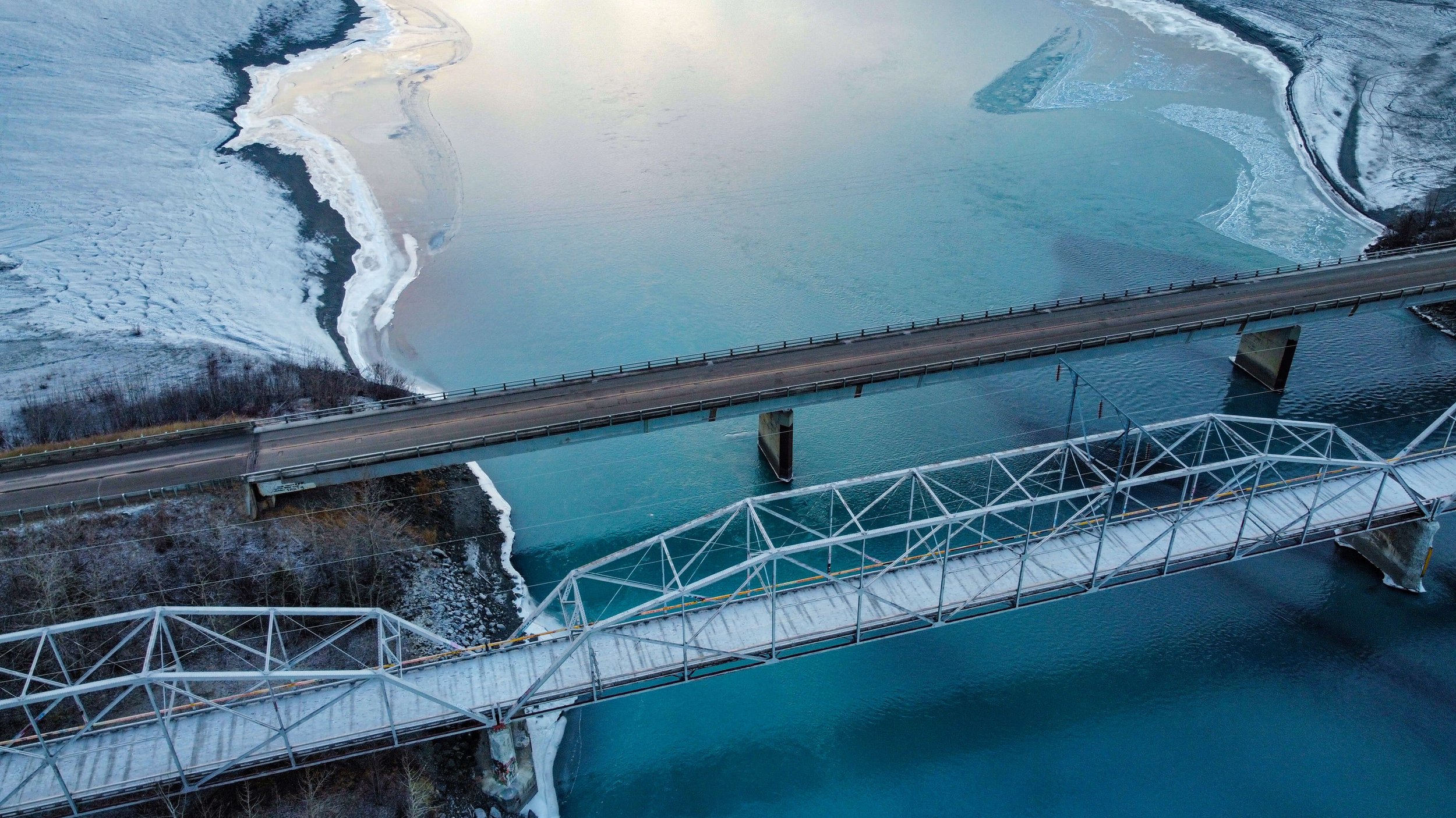 a drone photograph taken from the air of a road, highway bridge, train rail bridge, over partially frozen glacial blue river, taken in the Alaska wilderness, Alaska, USA, road shot, winter highway.