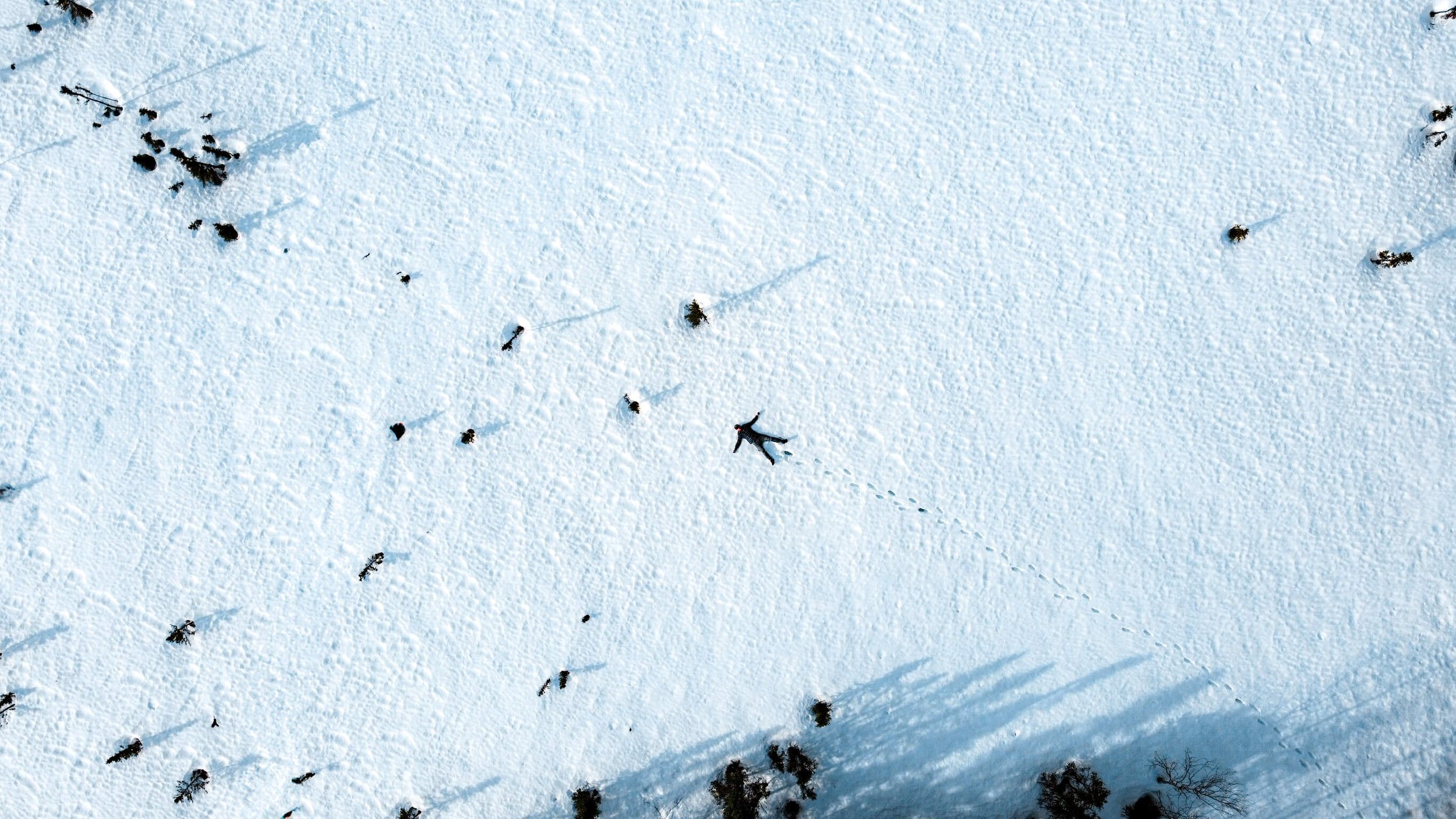 a drone photograph taken from high in the air of a lone male man laying down in the snow of the Alaska wilderness, Alaska, USA, snow boot prints, by himself, snow angel.