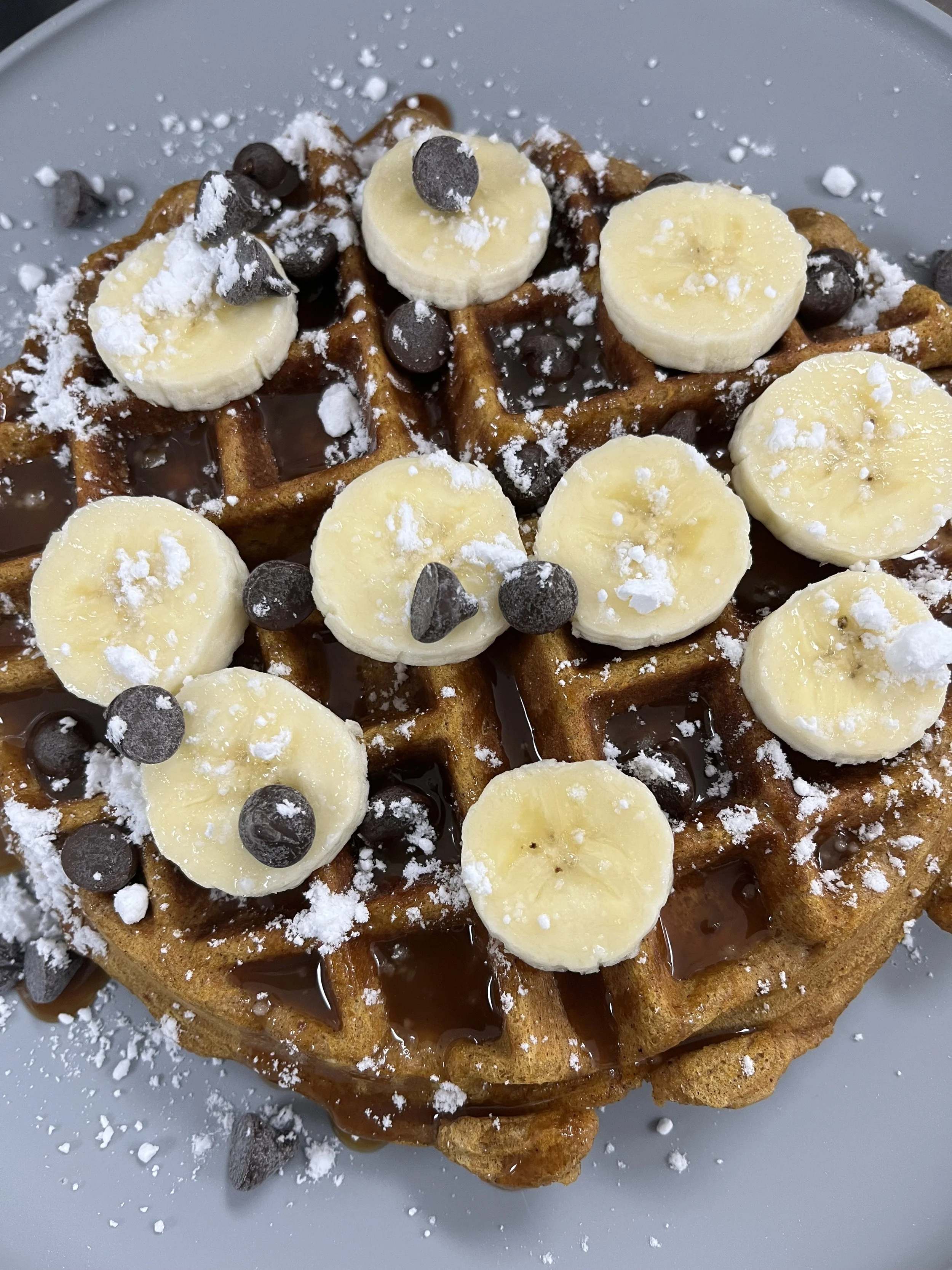 Waffle topped with banana slices, chocolate chips, powdered sugar, and drizzled with syrup.