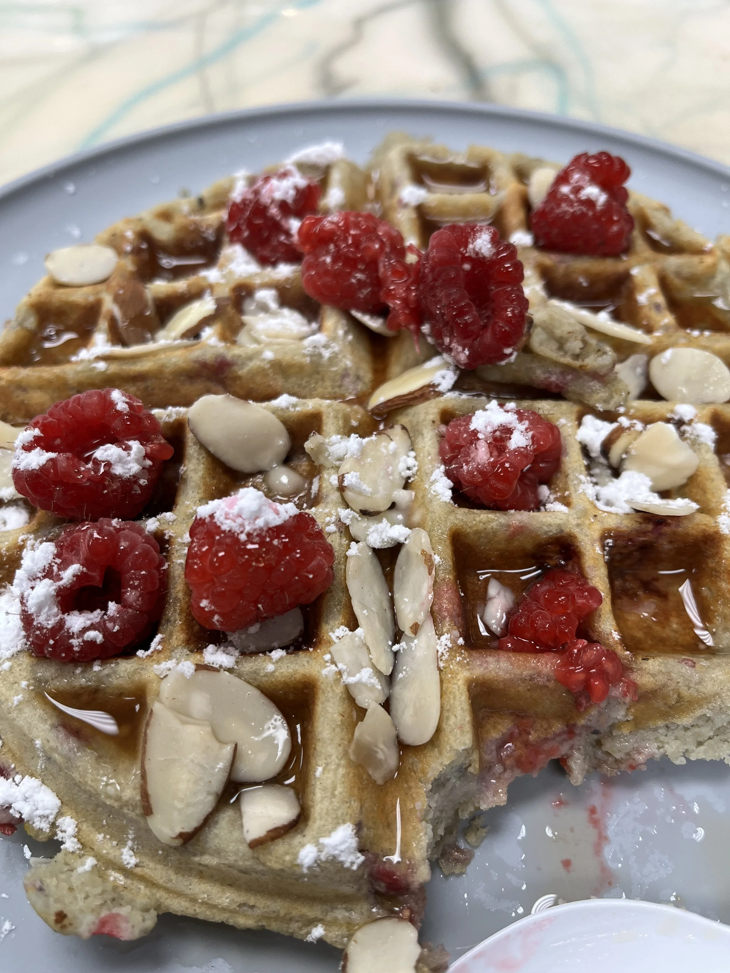 Close-up of a waffle topped with raspberries, sliced almonds, powdered sugar, and syrup on a light-colored plate.