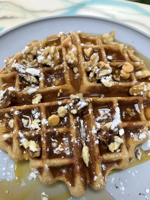 A golden brown waffle topped with crushed nuts, syrup, and powdered sugar on a white plate.