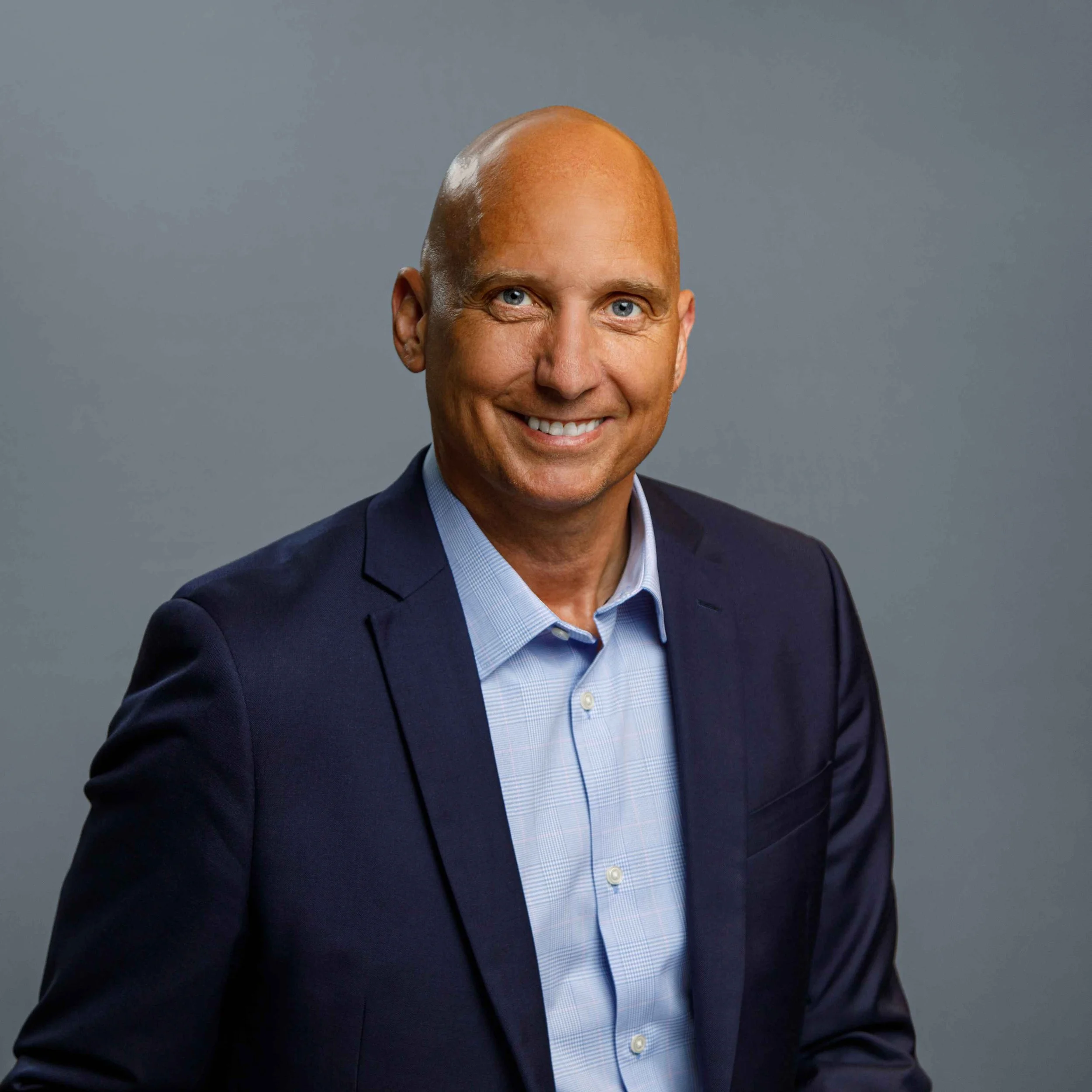 Headshot of a bald man wearing a navy blue suit and light blue shirt, smiling against a gray background.