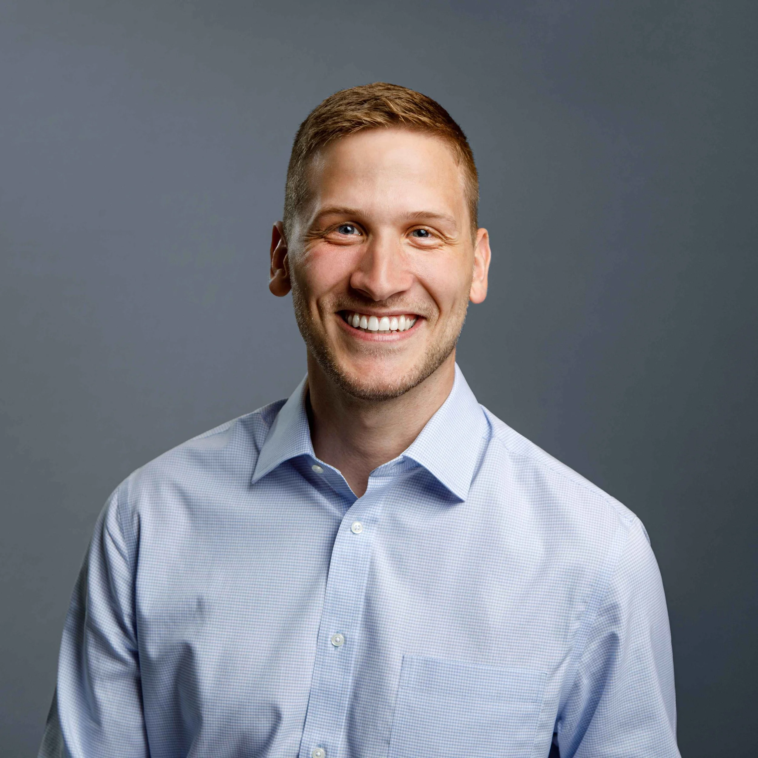 A smiling young man with short light brown hair, wearing a light blue button-up shirt, standing against a gray background.