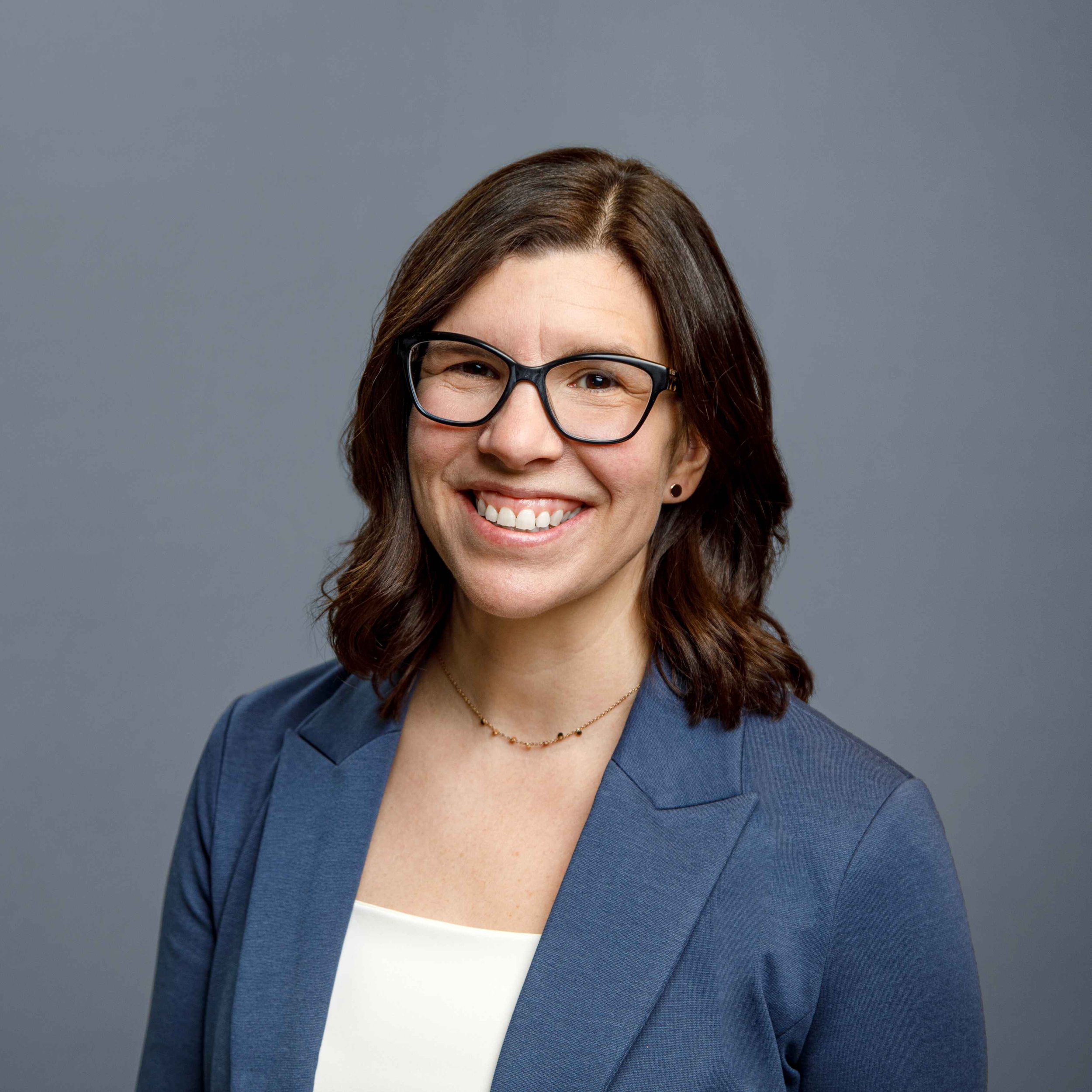 Smiling woman with brown hair, glasses, wearing a blue blazer and white top, against a plain gray background.
