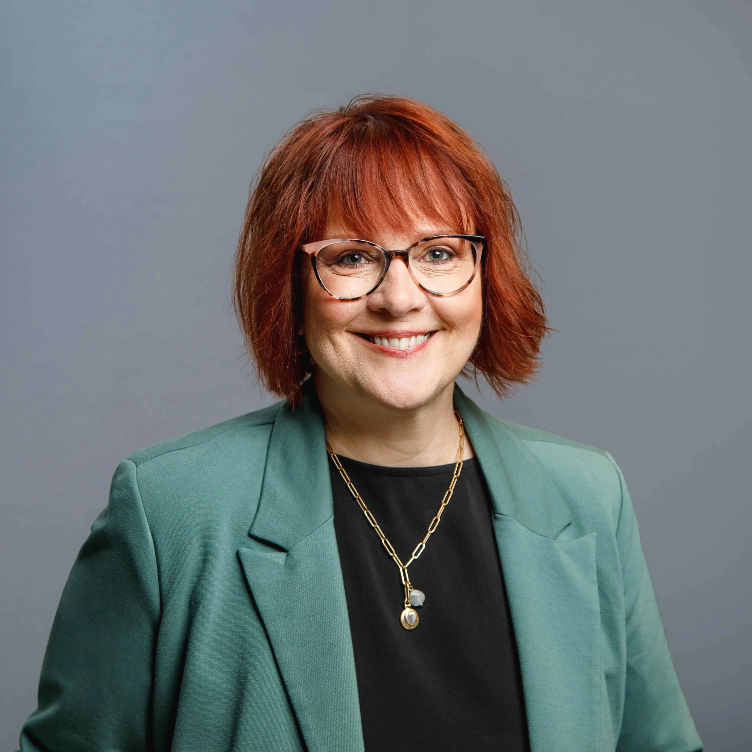A woman with red hair, glasses, and a green blazer smiling at the camera against a gray background.