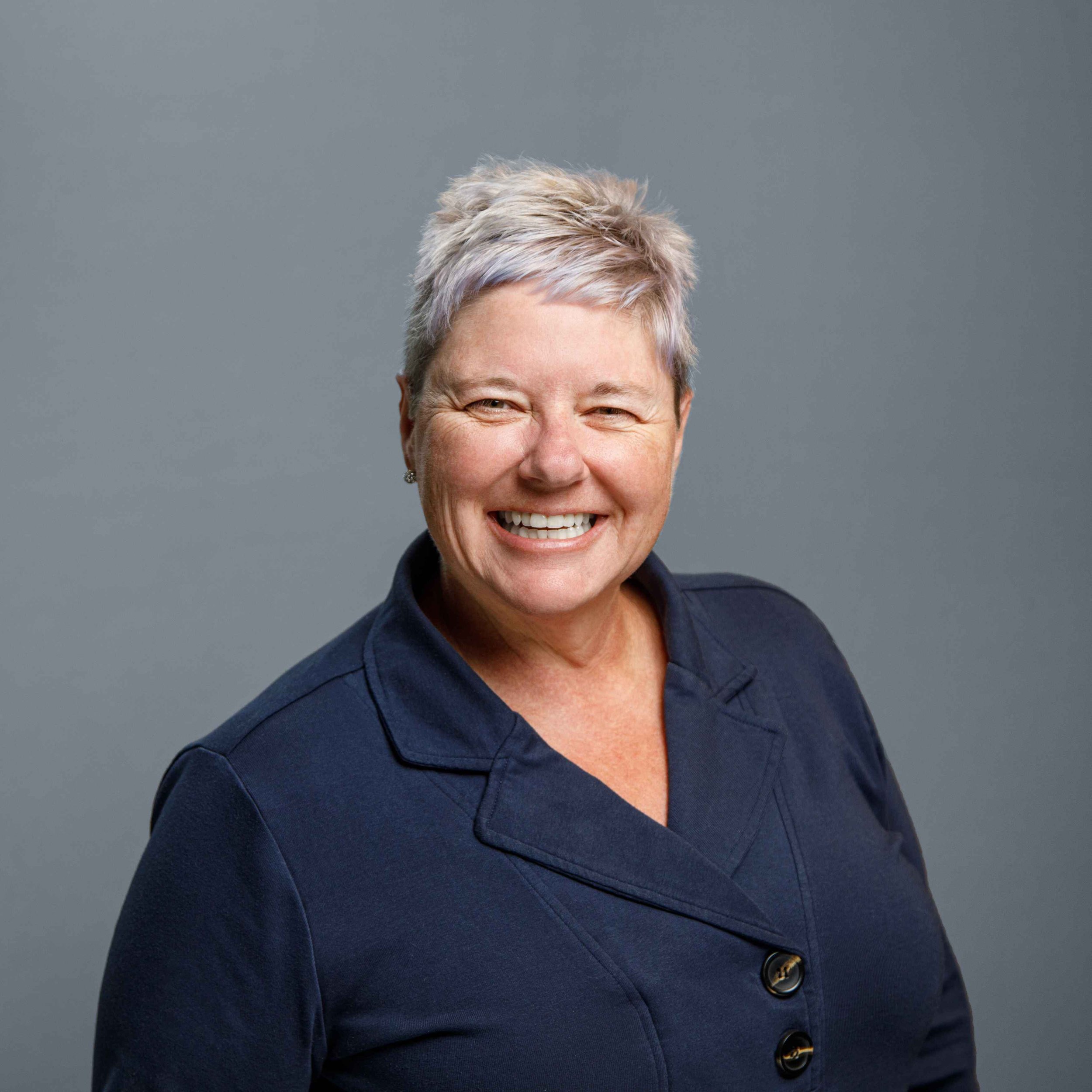 Smiling woman with short gray hair, wearing a navy blue blazer, against a gray background.