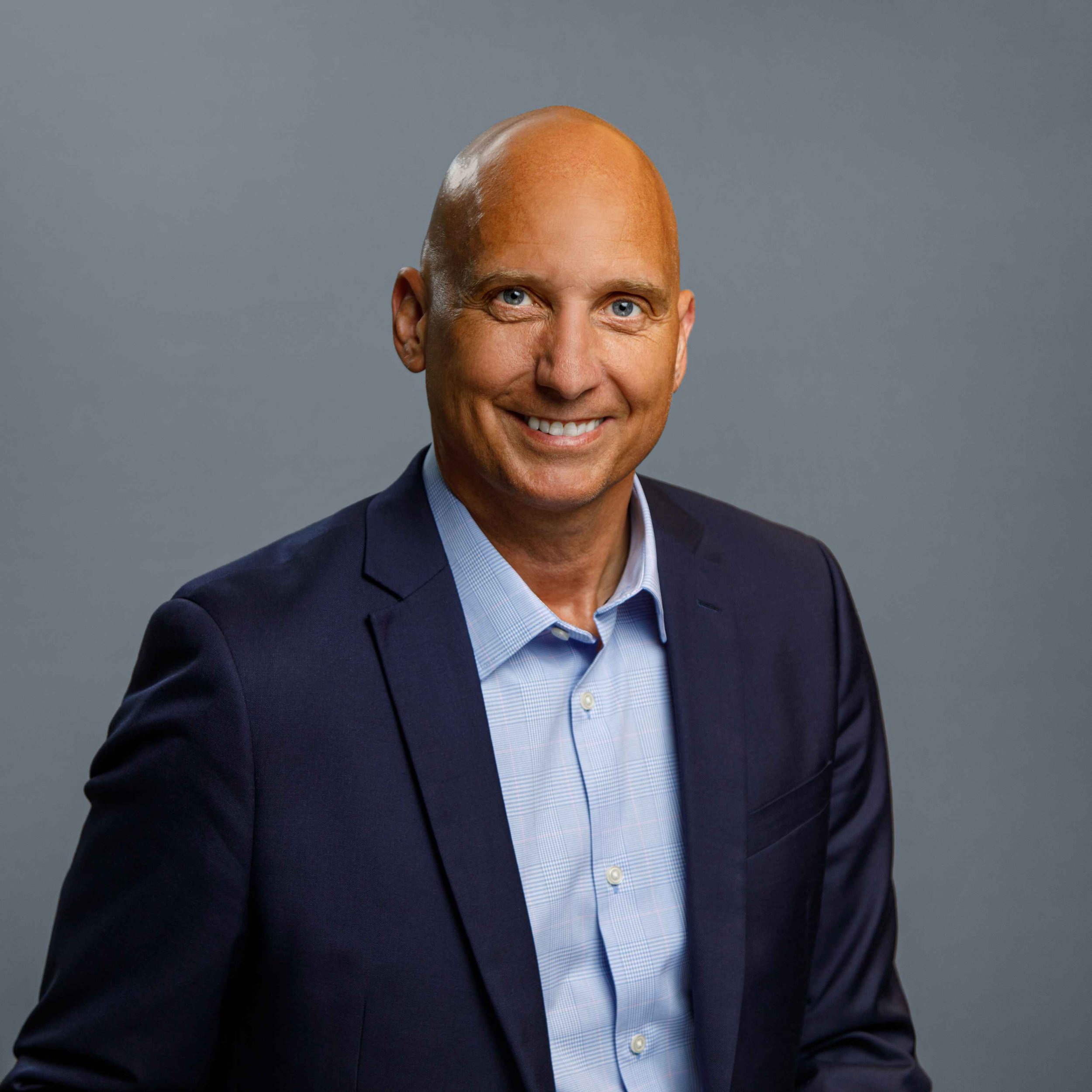 A smiling man with a bald head, wearing a navy blue suit jacket and a light blue dress shirt against a plain gray background.