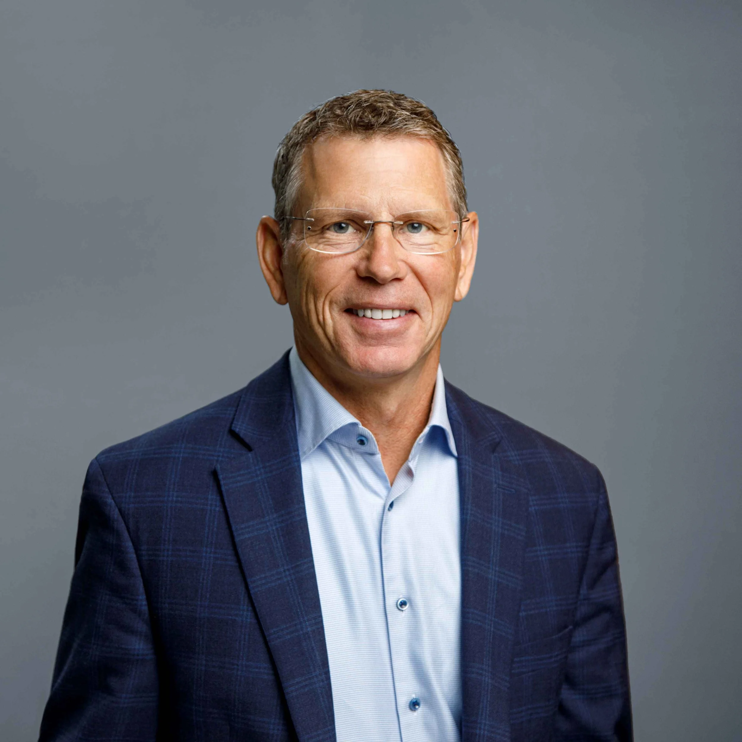 A professional man in a navy checkered blazer and light blue dress shirt, smiling against a plain gray background.