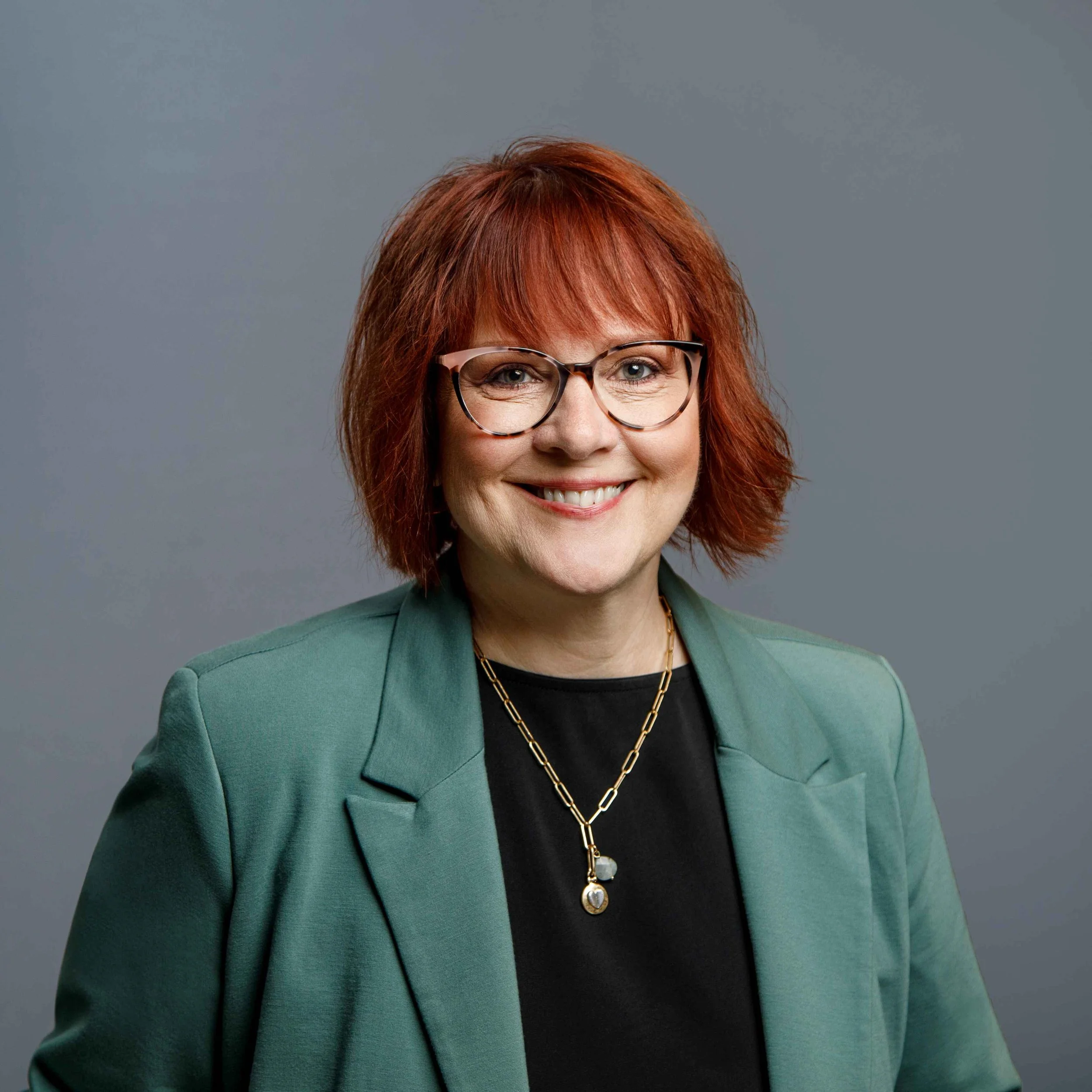 Professional woman with red hair, wearing glasses, a green blazer, and jewelry, smiling against a gray background.