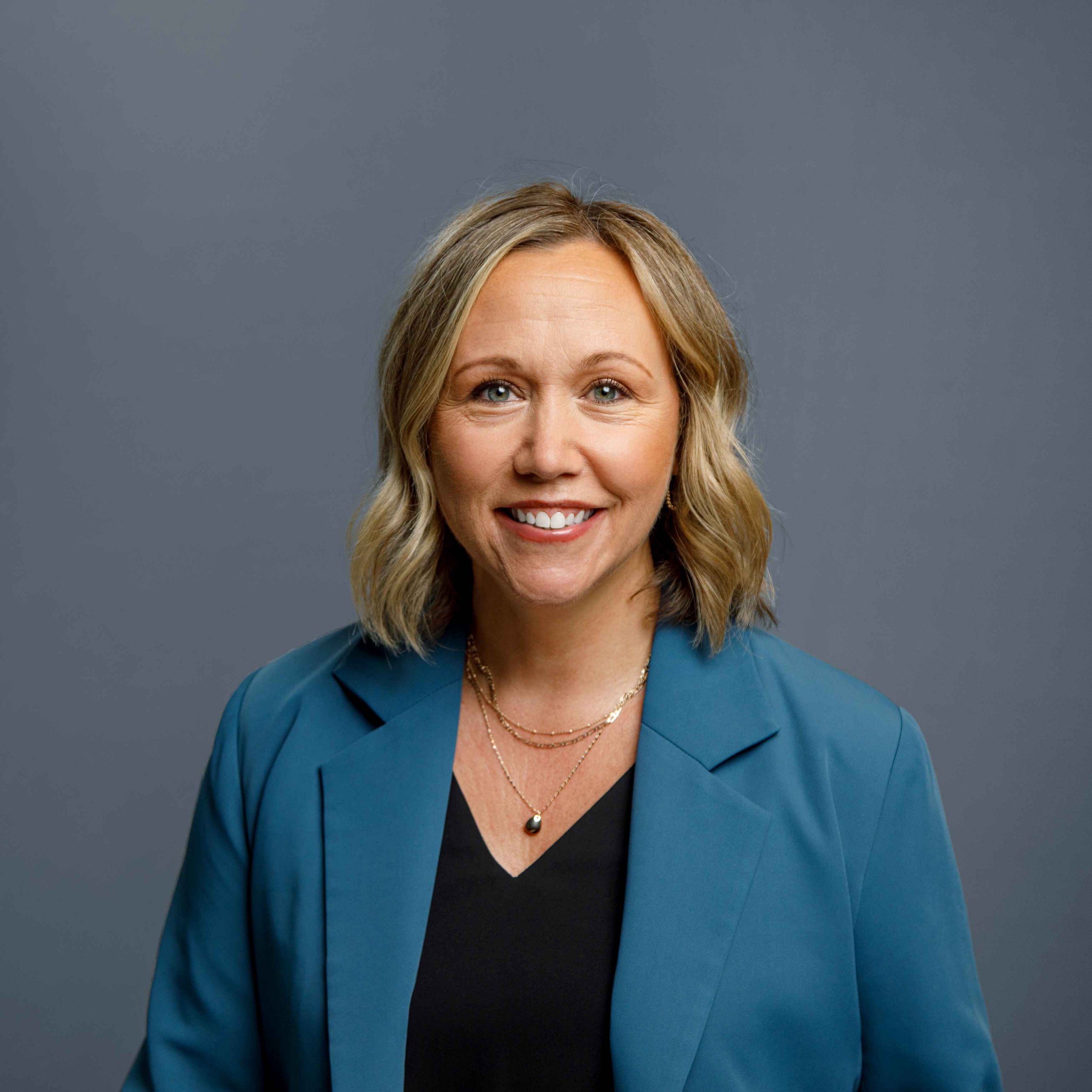 Portrait of a smiling woman with shoulder-length blonde hair, wearing a blue blazer, black top, and layered gold necklaces, against a grey background.