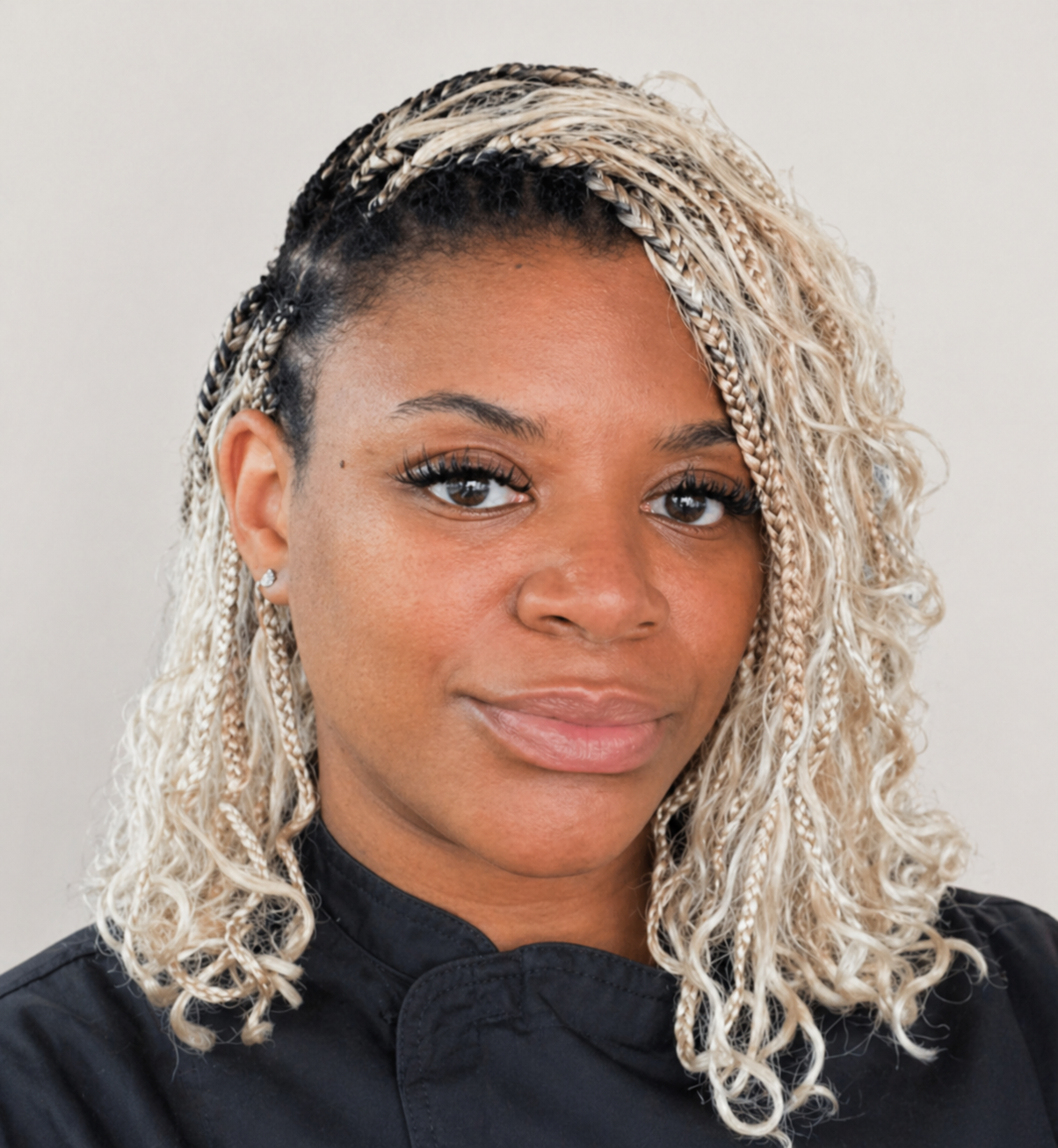 Close-up of a woman with blonde and black braided hair, wearing a black chef's coat, looking at the camera with a neutral expression.
