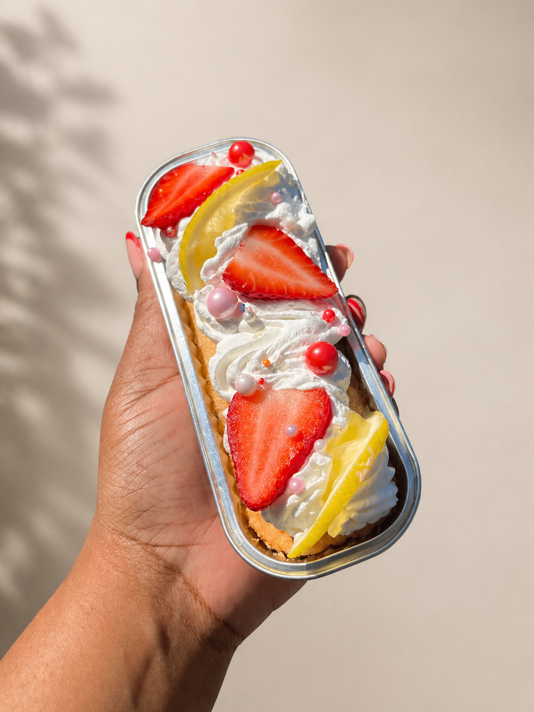 Hand holding a rectangular container with a simple cake topped with whipped cream, sliced strawberries, lemon slices, and colorful edible beads, against a plain background.