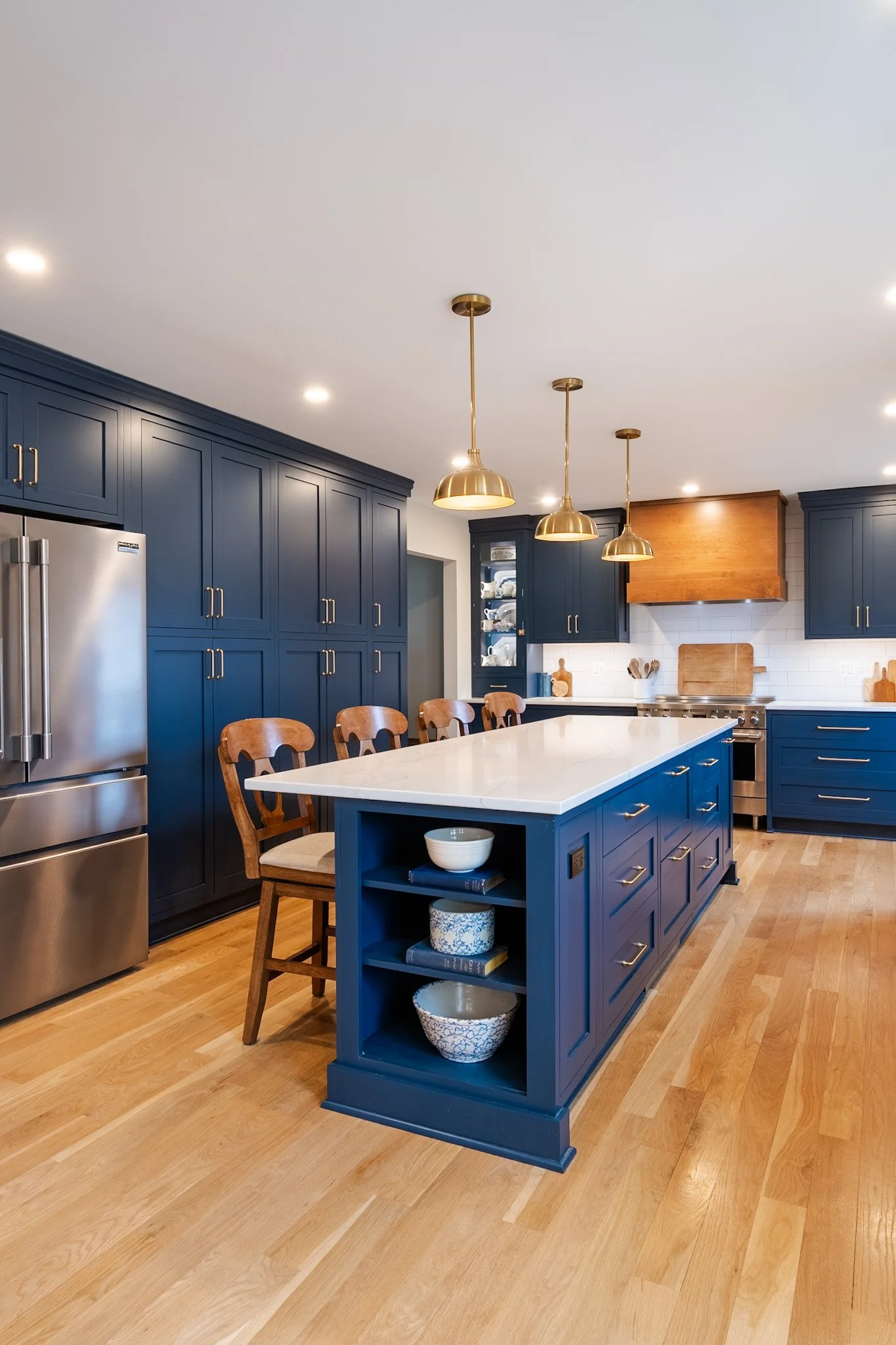 A modern kitchen with navy blue cabinets and a large kitchen island with a white countertop. The island has open shelves with bowls and books, and there are wooden chairs around it. Golden pendant lights hang above the island, and the kitchen features a stainless steel refrigerator, stove, and wooden flooring.