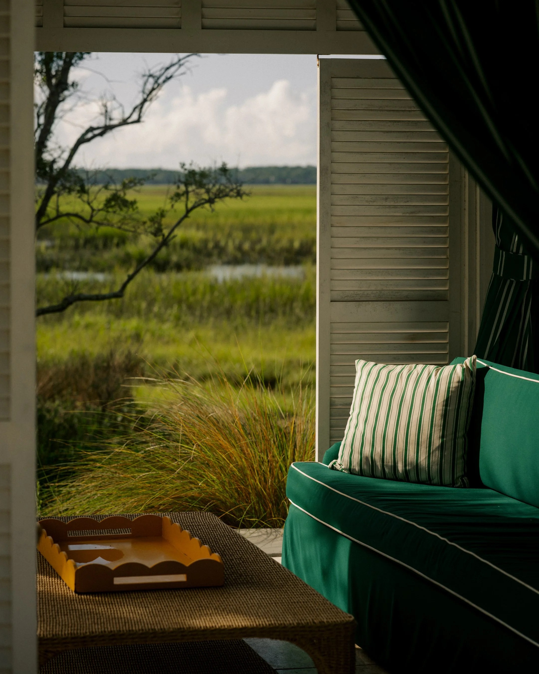 A view of a grassy field and trees seen through an open window of a porch or outdoor room, with a wicker table and a green cushioned chair with striped pillows in the foreground taken at The Dunlin.