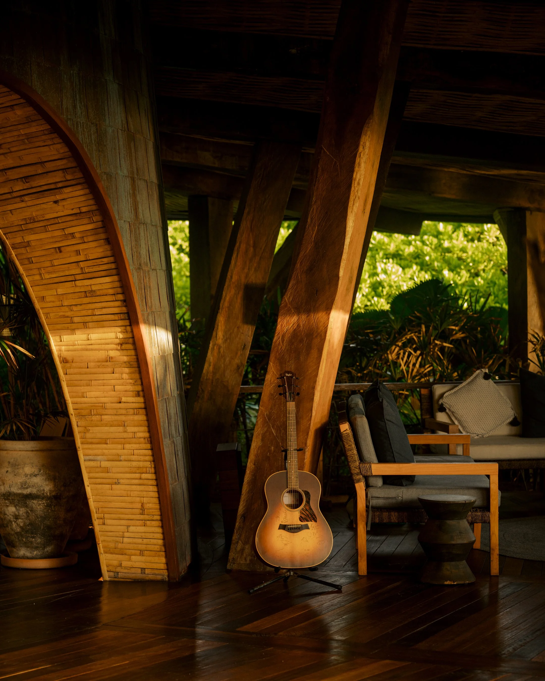 A wooden guitar standing on a stand, positioned in front of a wooden beam and a curved wooden structure, with a sofa and pillows in the background, and lush green plants visible outside. Photo taken a Naviva.