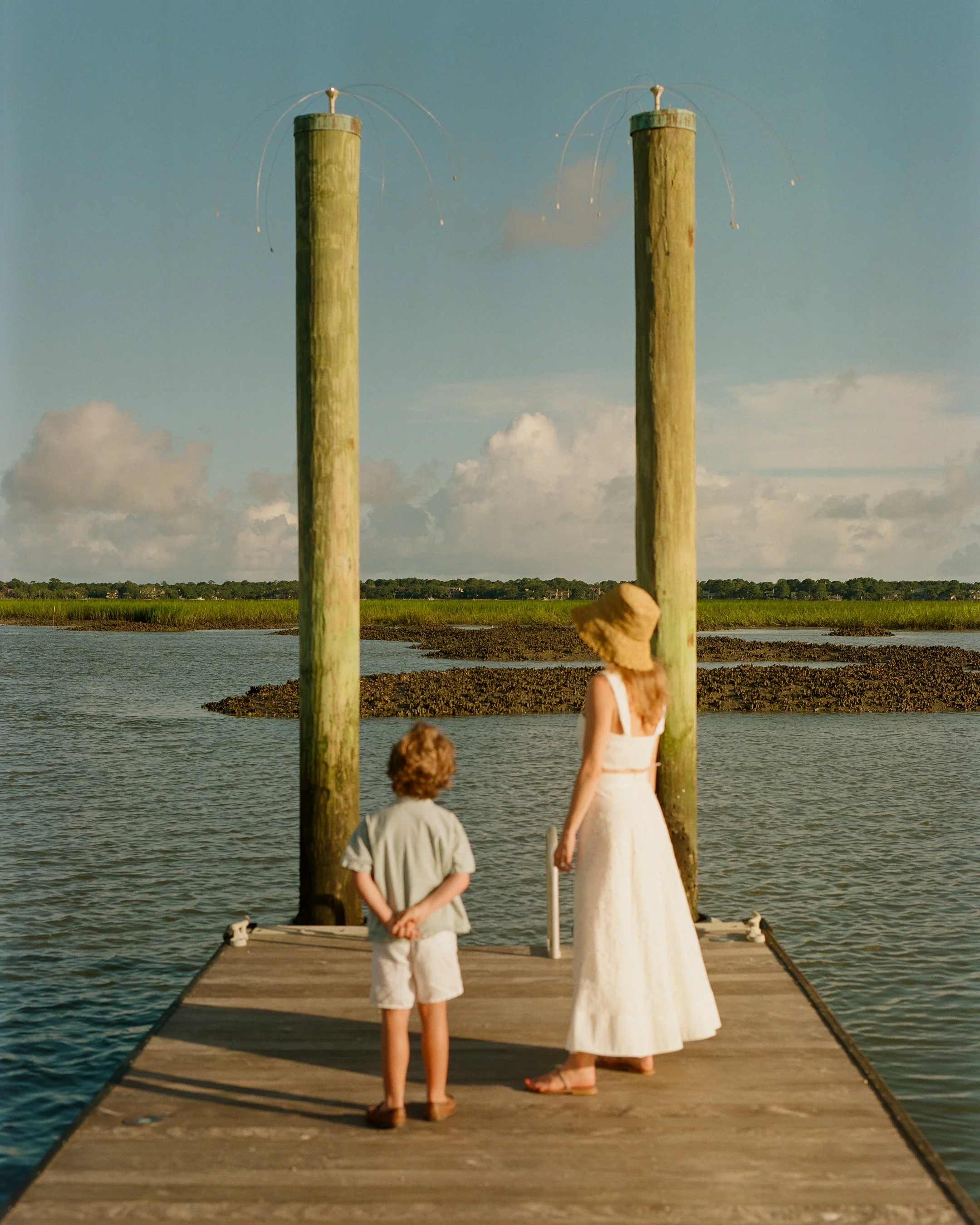 A woman and a young boy standing on a wooden dock by the water, with two tall green wooden posts with wire loops on top, under a partly cloudy sky. Photo taken at The Dunlin, Auberge Collection. 